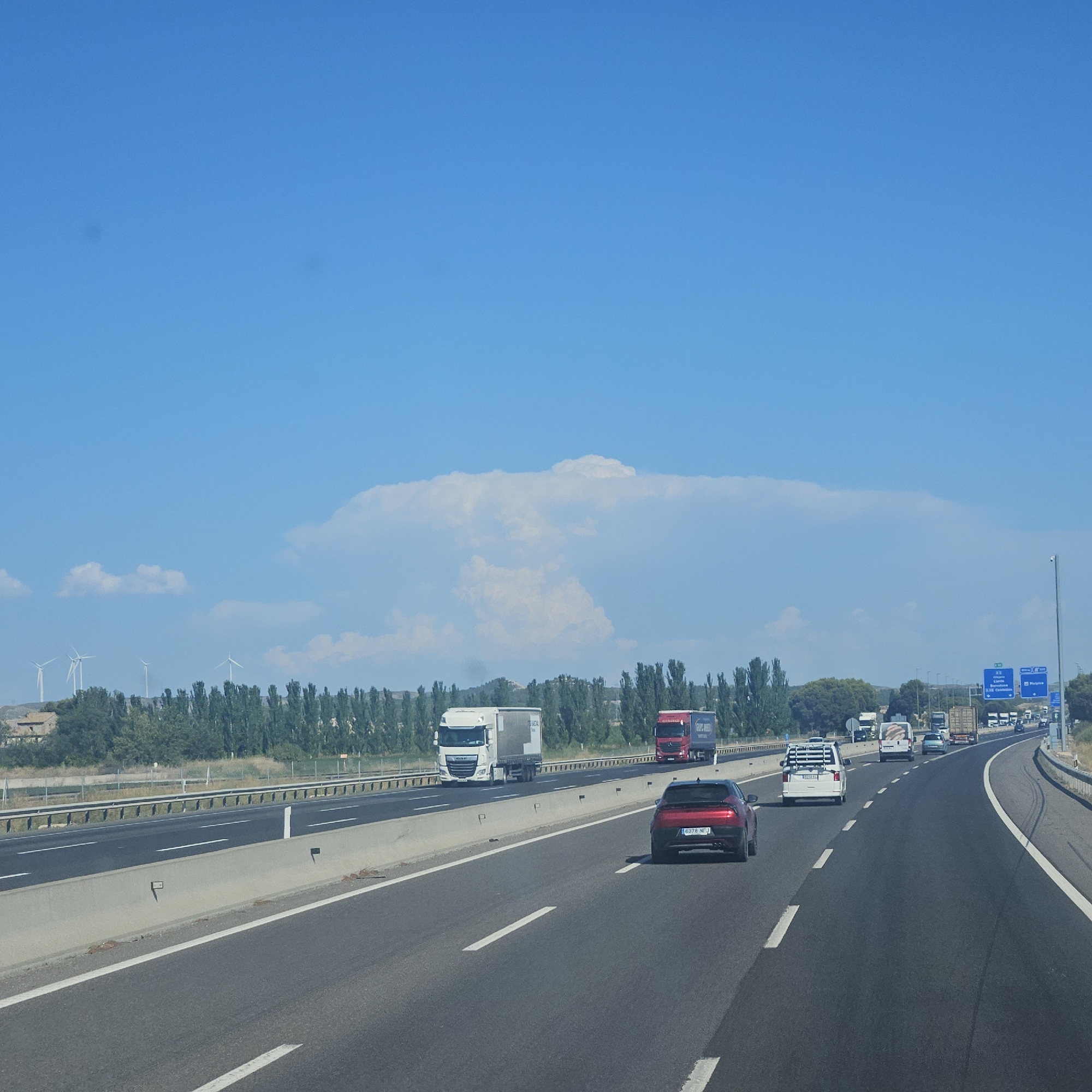 This is a daytime, eye-level, medium shot of a multi-lane highway in Villamayor de Gállego, Spain. The highway stretches into the distance, curving slightly to the right. The sky is clear blue with a large, distinct cumulonimbus cloud formation in the distance. A line of tall, slender green trees borders the highway on the left, with wind turbines visible behind them. Several vehicles are traveling on the highway, including a red sports car in the foreground, a white van, and multiple trucks. Blue highway signs indicating directions to cities like Barcelona and Castellón are visible in the background. The road surface is asphalt with white lane markings. A concrete barrier separates the lanes of traffic. The scene depicts typical highway traffic under a bright sky with an impressive cloud formation.