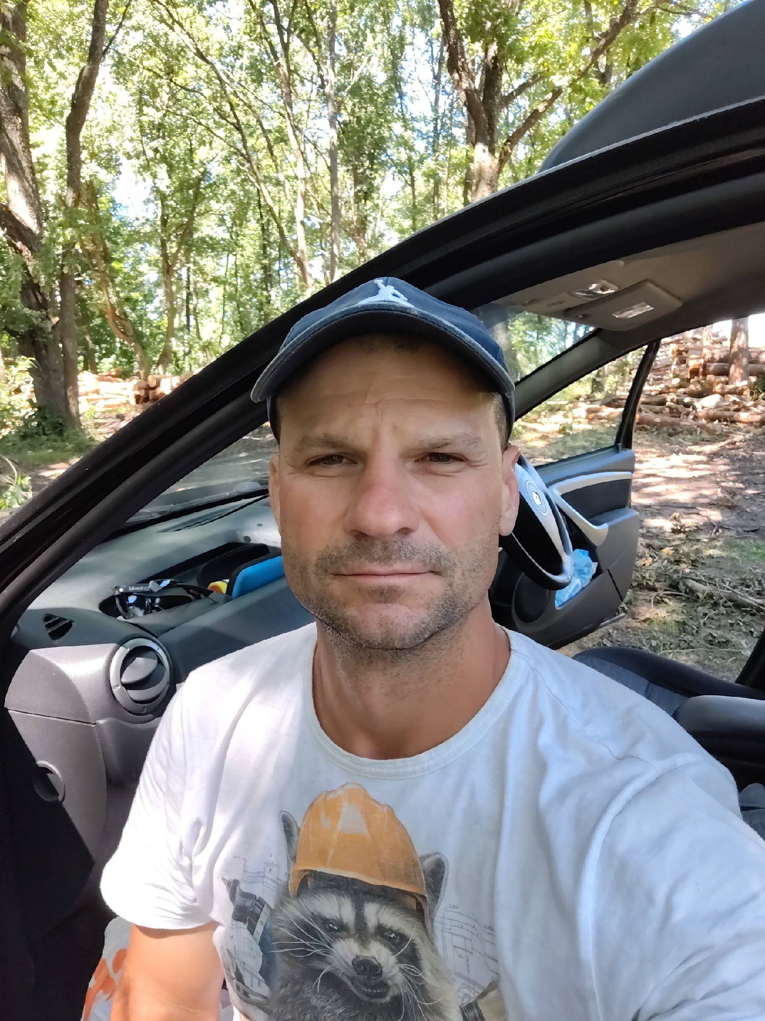 A man wearing a white t-shirt with a raccoon graphic and an orange hard hat, and a blue baseball cap, is sitting in the driver's seat of a car. The car's driver-side door and hood are open, revealing the interior. The background shows a wooded area with trees and a pile of logs. The car appears to be parked off-road, on a dirt surface. This scene is located within the Бабовіцкі сельскі Савет, Belarus.