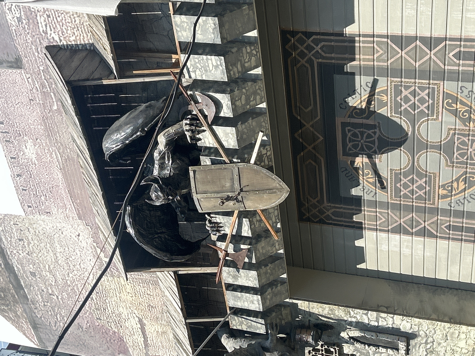 The image shows a close-up of a building's facade, featuring architectural elements reminiscent of a medieval castle. A large, dark, dragon-like sculpture with outstretched wings and claws is prominently displayed, holding a shield. Two axes are affixed to the wall beneath the sculpture. The building's exterior exhibits a rough, stone-like texture on the upper portion, transitioning to a corrugated metal surface on the lower section. This lower section is decorated with a large, ornate graphic featuring geometric patterns and what appear to be Celtic knot designs. The scene suggests a themed establishment, possibly an entertainment venue or restaurant, designed to evoke a fantasy or historical atmosphere. The lighting indicates it is daytime, with strong shadows cast by the architecture and sculpture. There are no people visible in the image. This building is located in the city of Perm, Russia, specifically at 33 Komsomolsky Prospekt, which houses the "Celtic Moon" pub.