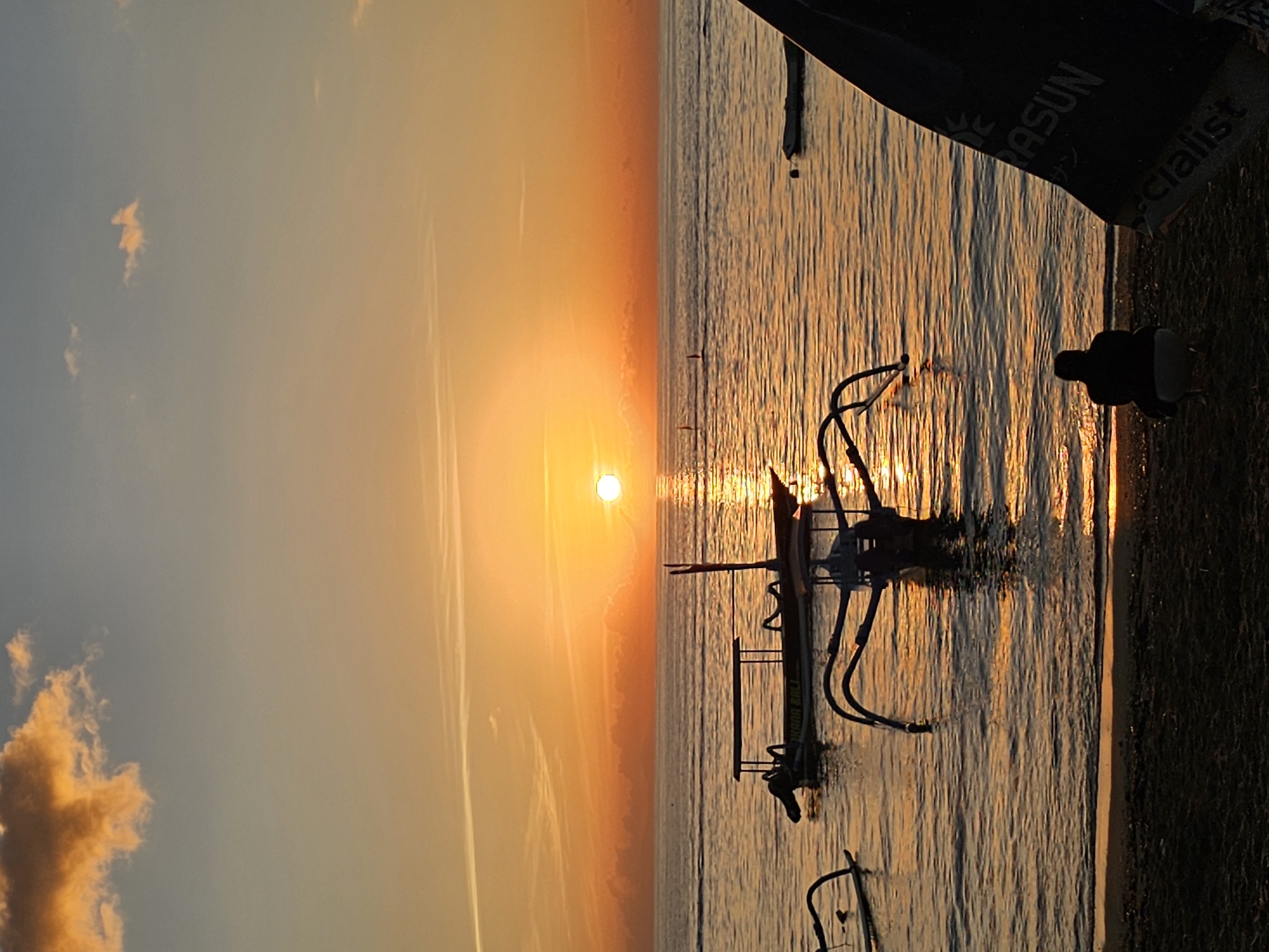 The image depicts a sunset over water, likely a beach scene in Denpasar, Indonesia. A small outrigger boat is partially submerged in the water, its silhouette highlighted by the setting sun. The water reflects the golden light. Part of a black object (likely a boat or surfing equipment) is visible in the foreground. The sky transitions from blue to orange hues, with faint clouds in the corner. Contrails are also seen in the sky. There are no people visible in the scene.