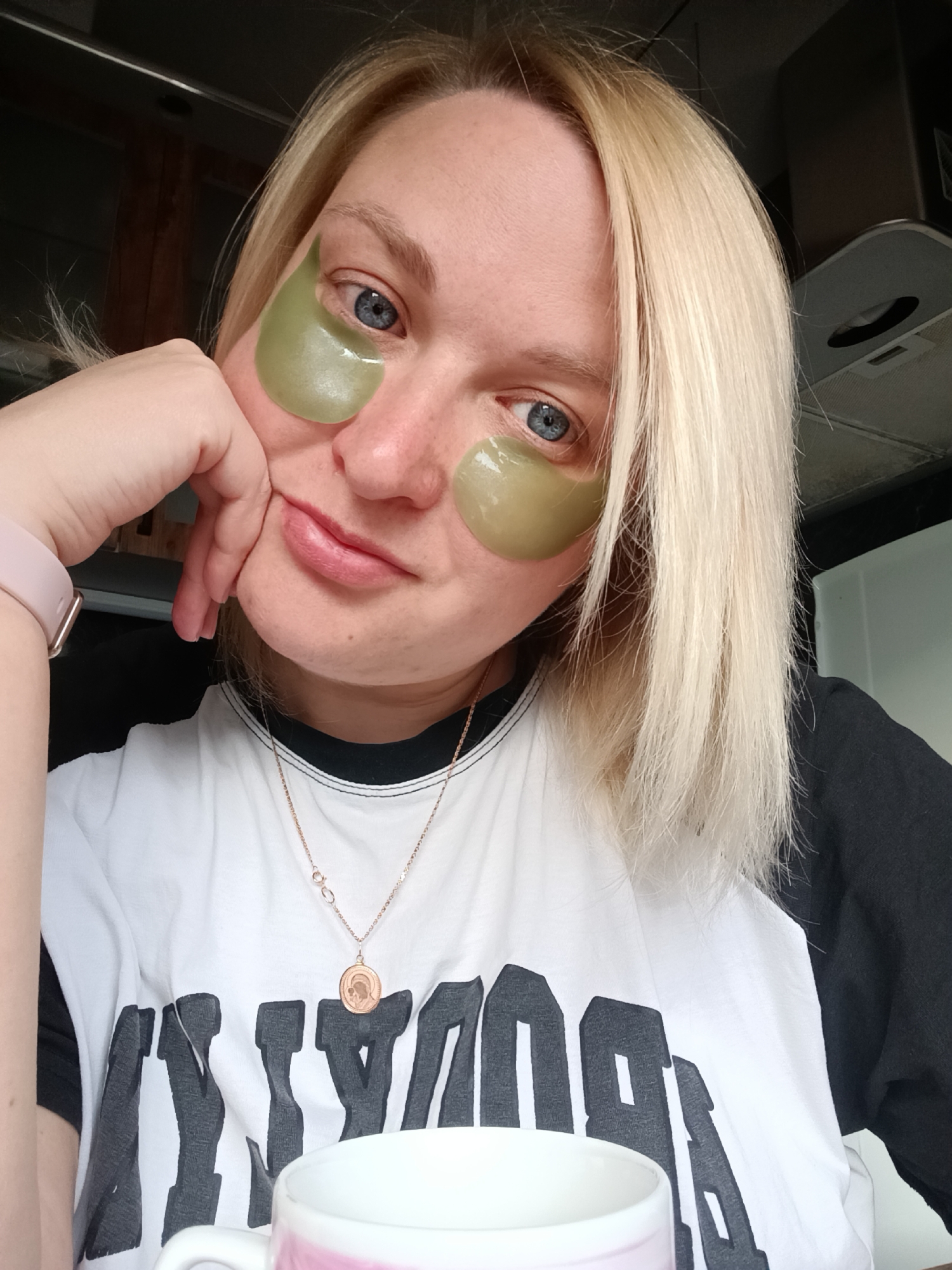 A woman with blonde hair and blue eyes poses indoors, with green under-eye patches on her face. She is wearing a black and white baseball t-shirt and a gold necklace with a pendant. Her left hand rests on her cheek, supporting her head. A pink watch is visible on her left wrist. In the foreground, a white mug with a pink base is partially visible. The background includes a kitchen with a dark countertop and light-colored walls, with an appliance visible at the top right. The photo was taken in the city of Chaikovsky, Russia.