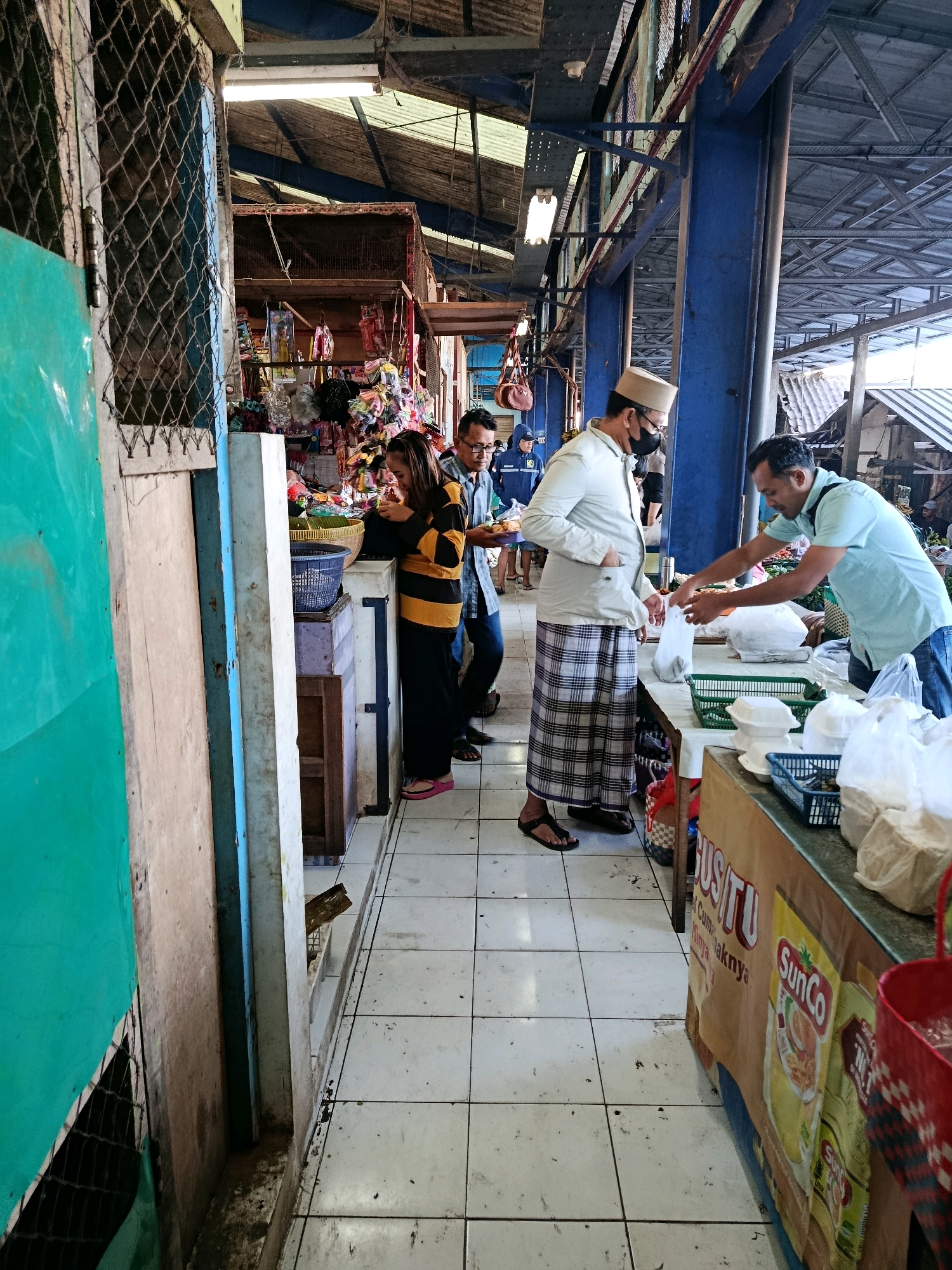 The image depicts an indoor market scene in Bululawang, Indonesia. In the foreground, a man in a light-colored shirt, a tan cap, and a plaid sarong is being served by another man in a light blue shirt who is placing items into a plastic bag. Both men are standing at a stall. To the left, a woman wearing a yellow and black striped shirt and black pants is looking down at something she is holding. Further to the left, another man in a gray shirt is visible. The market stall features various items for sale, including produce, packaged goods, and baskets. The environment appears to be an open-air or semi-covered market with a tiled floor and a corrugated roof supported by blue metal beams. The overall scene is one of a busy market with people engaged in commerce.