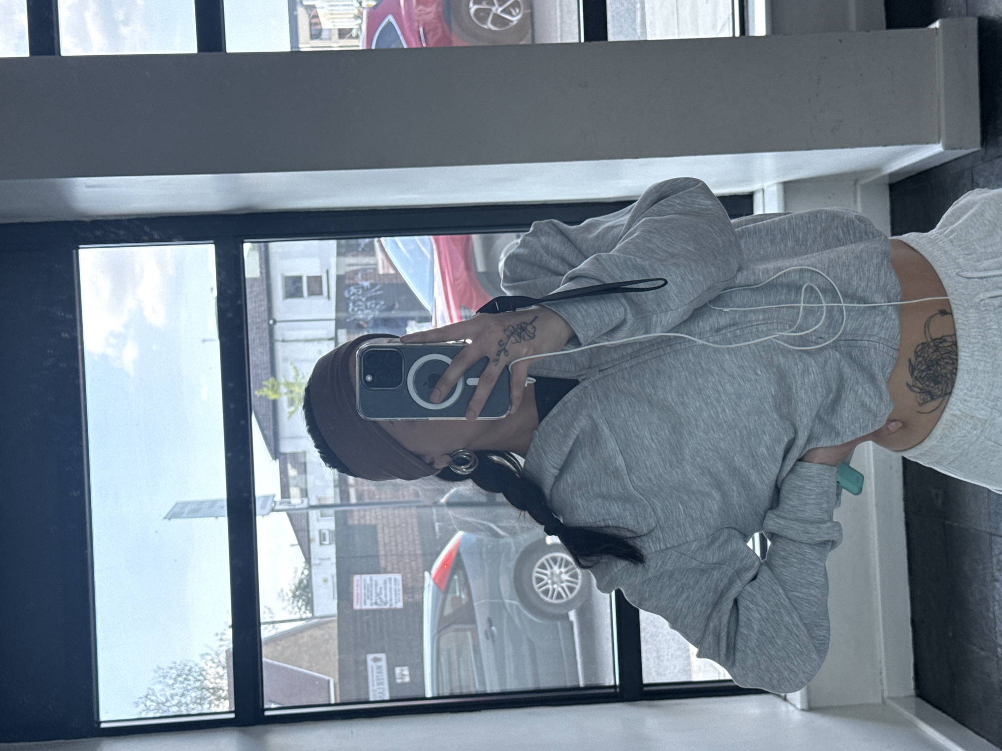 The image captures a mirror selfie of a person standing indoors, reflected in a large window or glass partition. The subject is seen from the chest down, wearing a dark headband and their dark hair in a braid. They are dressed in a grey hooded sweatshirt and light grey sweatpants, indicative of casual attire.
The person holds a smartphone, likely an iPhone with a MagSafe ring visible on its clear case, in their left hand to take the photo. A small, stylized floral tattoo is visible on the back of their left hand. White wired earphones are draped around their midsection. A larger, abstract tattoo is partially visible on their lower back/torso, just above the waistband of their sweatpants. A small, light green object is held in their right hand. The subject also wears circular earrings.
The reflection in the window reveals an outdoor urban or suburban environment during daytime, with a clear sky and some clouds. Visible elements include buildings, parked cars (one red, one dark grey hatchback), and hints of trees or foliage. On one of the reflected buildings, a sign with partially visible and reversed text, potentially "AJ K...AW," and smaller text below it, can be seen. The indoor setting features dark window frames and what appears to be a light-colored wall or floor. The scene suggests a casual moment in London, United Kingdom.