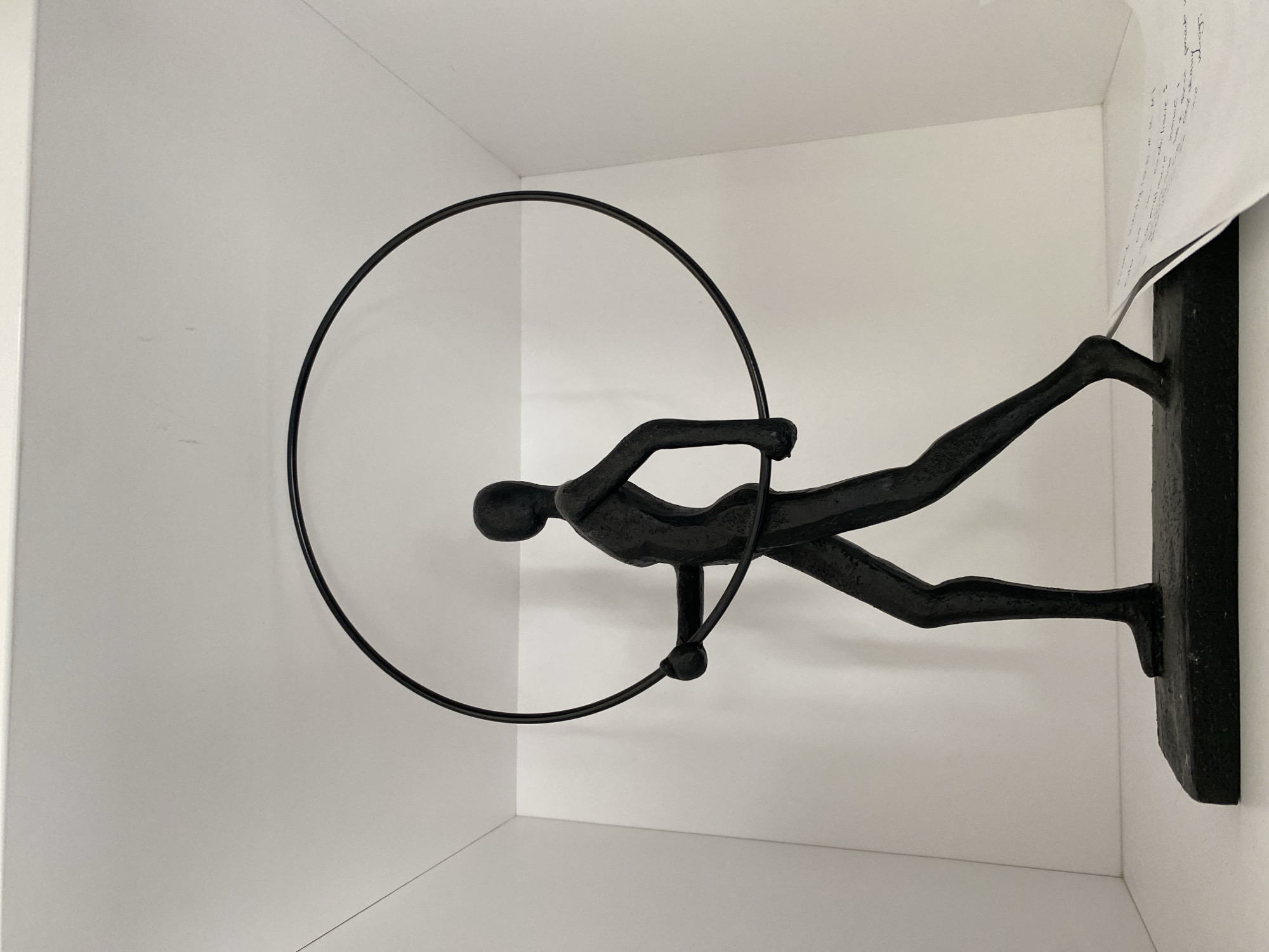 A black, stylized sculpture of a human figure holding a large hoop stands on a rectangular base. The figure is depicted in mid-stride, with one leg extended forward and the other bent. The hoop is circular and held horizontally with both hands. The sculpture is positioned within a white shelf or cubby. A piece of paper with handwritten text is visible on the right side of the base, partially obscuring the shelf. The background is a plain white wall. The scene is likely an indoor setting, such as a home or gallery, and the location in Buk, Poland, is not visually determinable from the image.