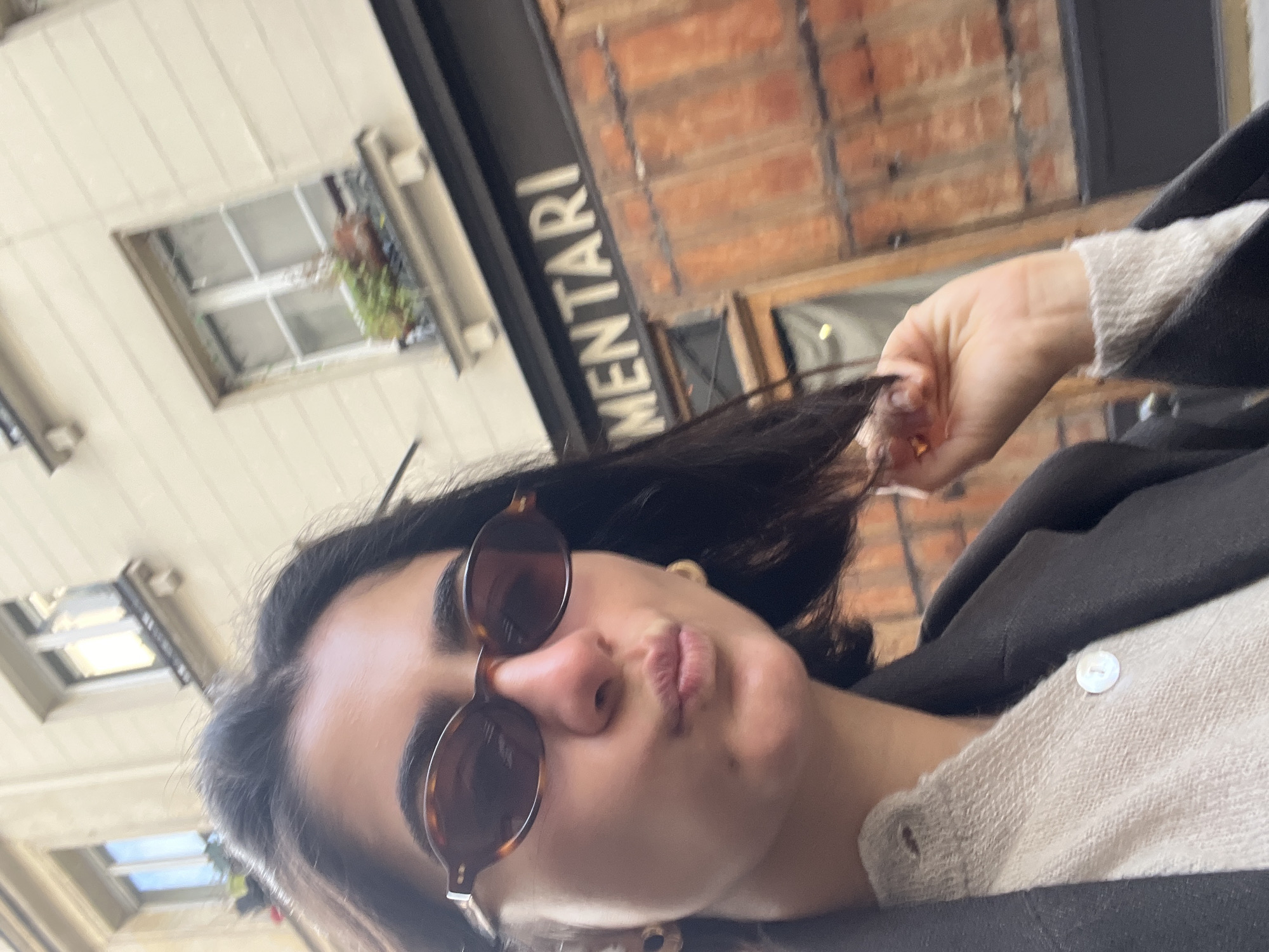 The image is a dynamic, angled close-up selfie of a young woman in an urban setting, consistent with Paris, France. She looks directly at the camera, pouting her lips in a "kissy face" or "duck face" expression and wearing brown tortoiseshell-patterned sunglasses that obscure her eyes. Her dark hair frames her face, and her right hand is visible near her ear, gently holding a strand of her hair. She is dressed in a light-colored, possibly cream-toned, textured top or cardigan with a white button, layered under a dark jacket or coat. A small gold-colored earring is also visible.
The background shows architectural elements: a light-colored building wall with a multi-paned window to the upper left, and a warm-toned brick building further to the right. A prominent dark sign with white capital letters spelling "MENTARI" is visible behind her head, though the initial part of the word is cropped out. The lighting suggests it is daytime. The overall mood is casual and playful.