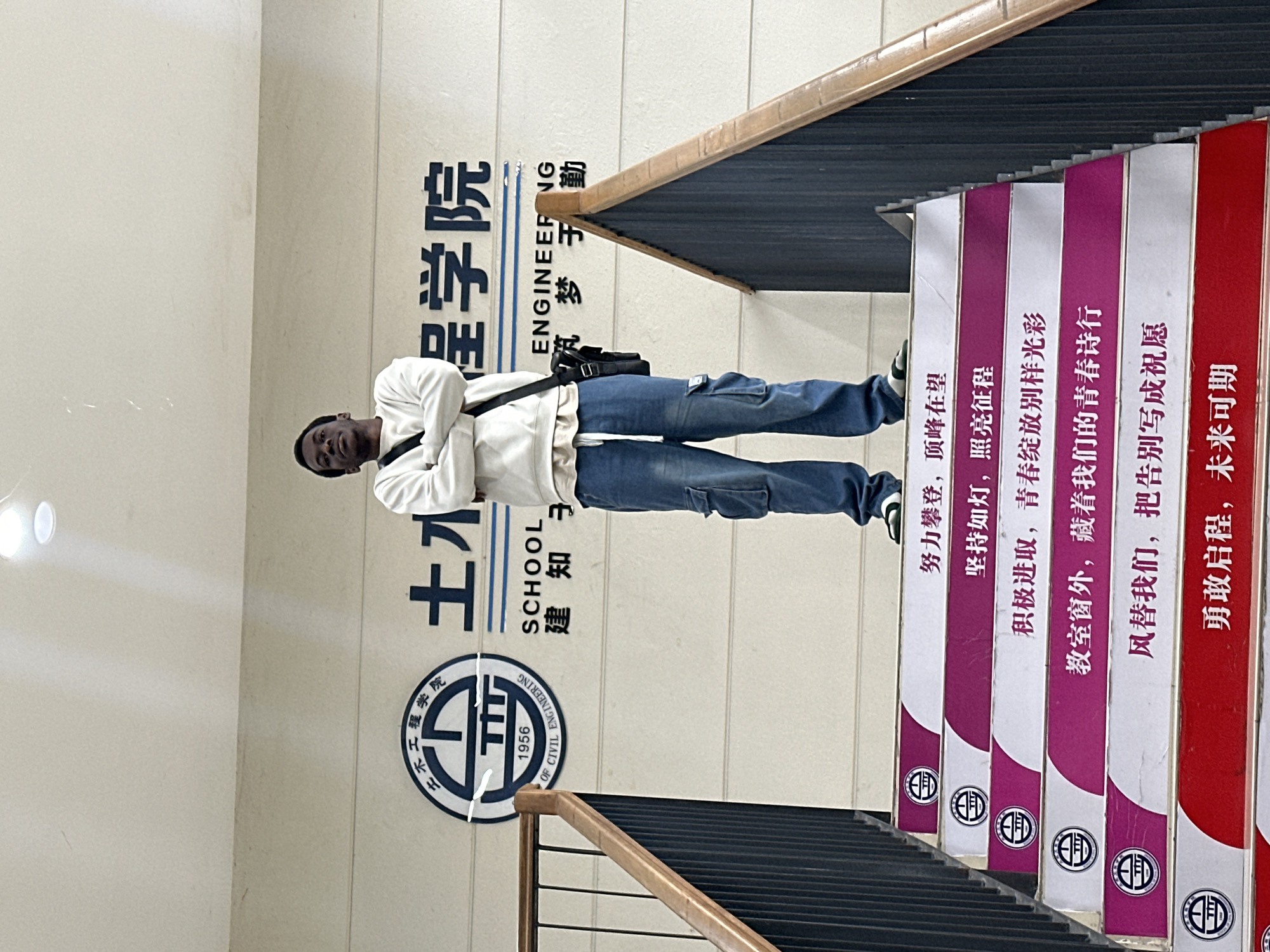 A man stands on a staircase in what appears to be an academic building. The wall behind him has Chinese characters and the words "SCHOOL OF CIVIL ENGINEERING 1956." Stair risers are painted with encouraging Chinese phrases in white on red and purple backgrounds. The man is wearing a white sweatshirt and blue jeans. This is the Jiangjin campus of the Chongqing University of Science and Technology.