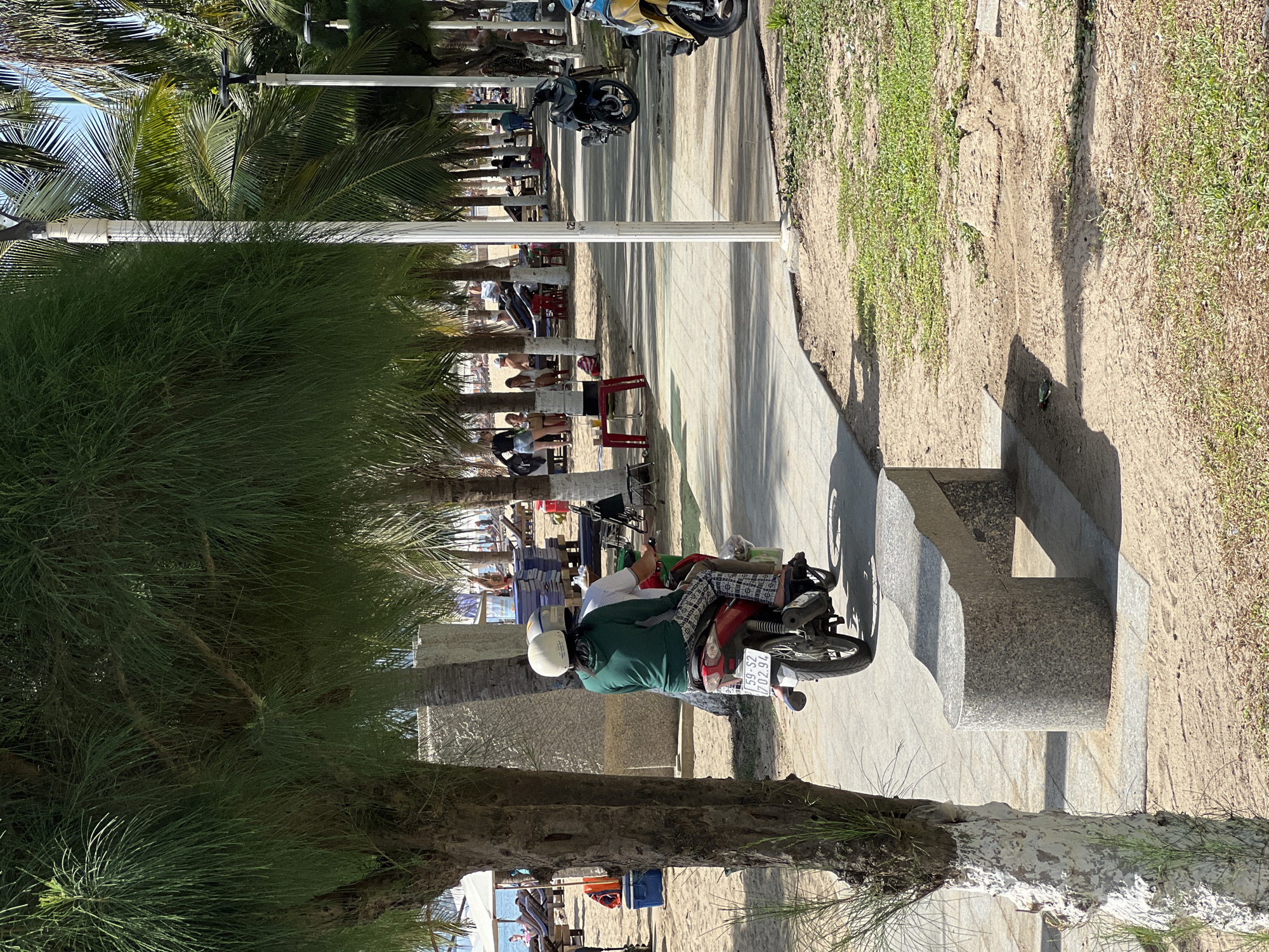 In Nha Trang, Vietnam, this image captures a street scene adjacent to a beach. A red scooter with two occupants, a driver and a passenger wearing helmets, is parked on a paved walkway. The driver, wearing a green top and patterned pants, is in focus. Behind the scooter, palm trees and other lush vegetation line the walkway. The sandy beach, with lounge chairs and umbrellas, is visible in the background, indicating a tourist area. The lighting suggests a bright, sunny day.