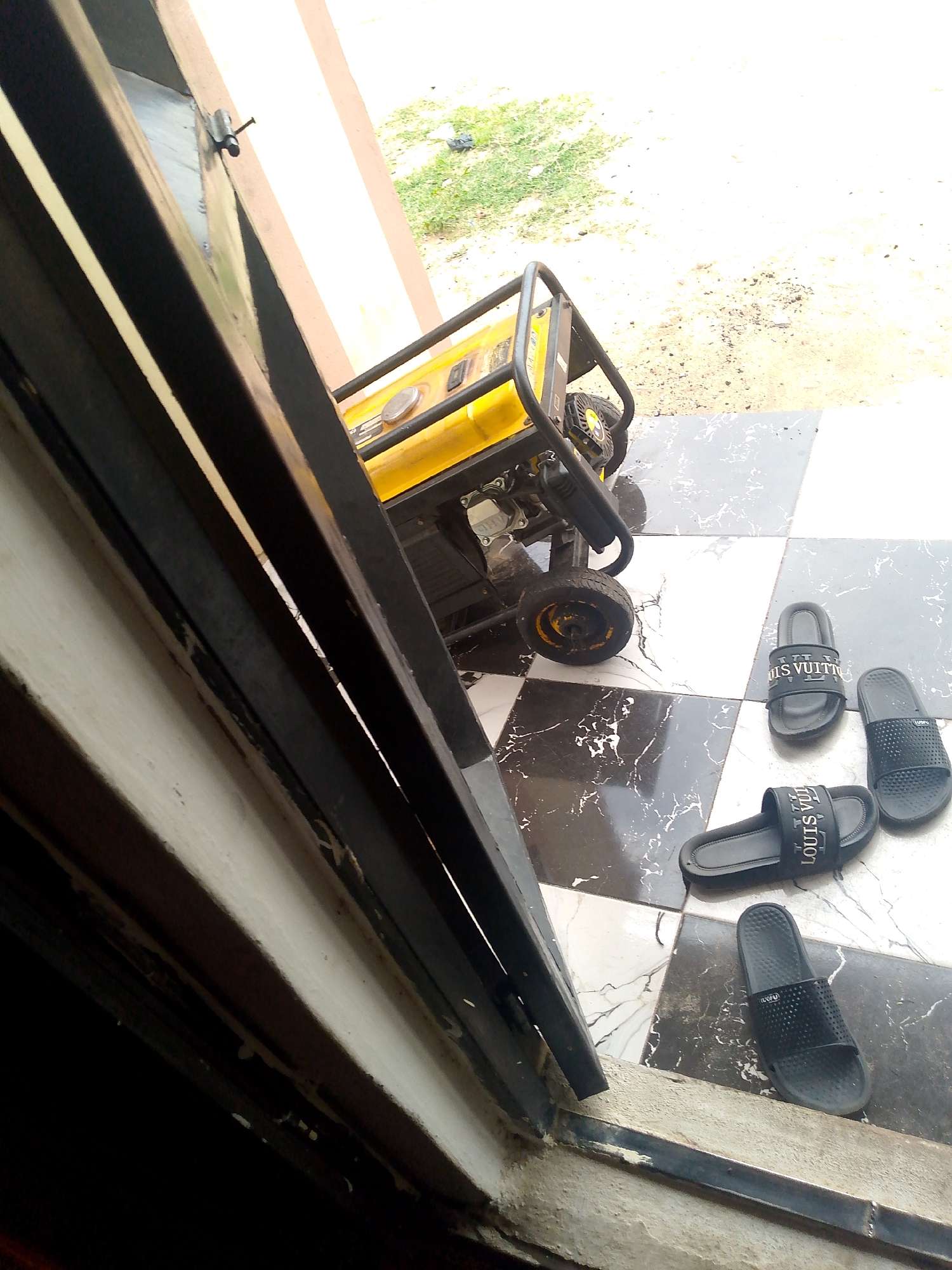 The image depicts a residential outdoor scene in Jauro Yinu, Nigeria. A yellow generator with a black frame and wheels is positioned on a tiled patio. Four black flip-flops are scattered on the tiles; two have the "LOUIS VUITTON" logo in white, and the other two are plain black. A partially open doorway with a black frame is visible on the left. The background shows unpaved ground with sparse green vegetation. The lighting suggests it is daytime, possibly overcast.
