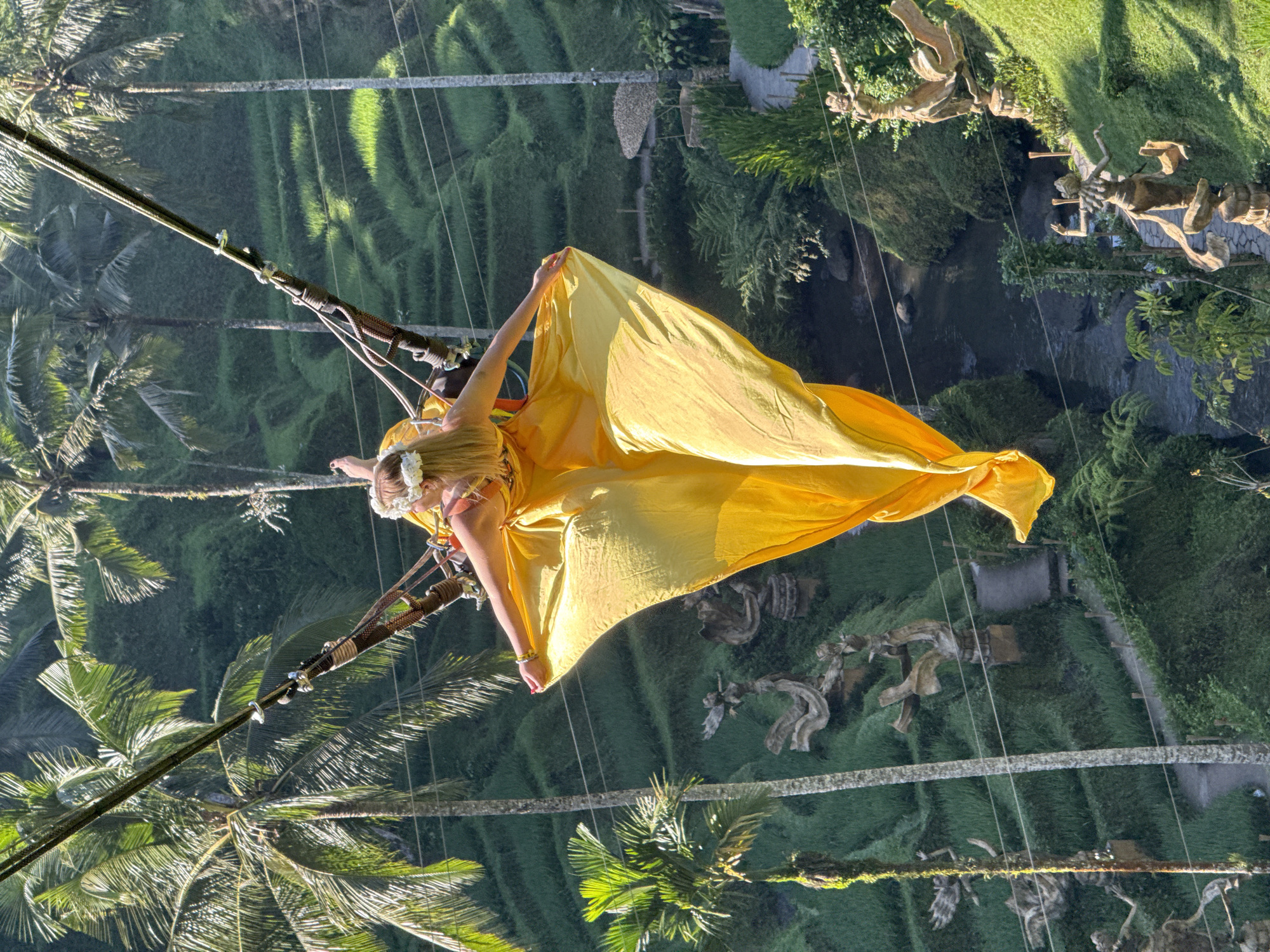 In Tegallalang, Indonesia, a woman is captured mid-swing on a high swing, her long, flowing yellow dress billowing around her. She is adorned with a white flower crown on her blonde hair. The swing is suspended by thick ropes against a backdrop of lush green terraced rice paddies and tall palm trees. Below, a winding path is visible, bordered by decorative statues of human figures. A small river flows through the gorge, adding to the scenic beauty of the location. The lighting suggests a sunny day, casting shadows and highlighting the vibrant colors of the landscape.