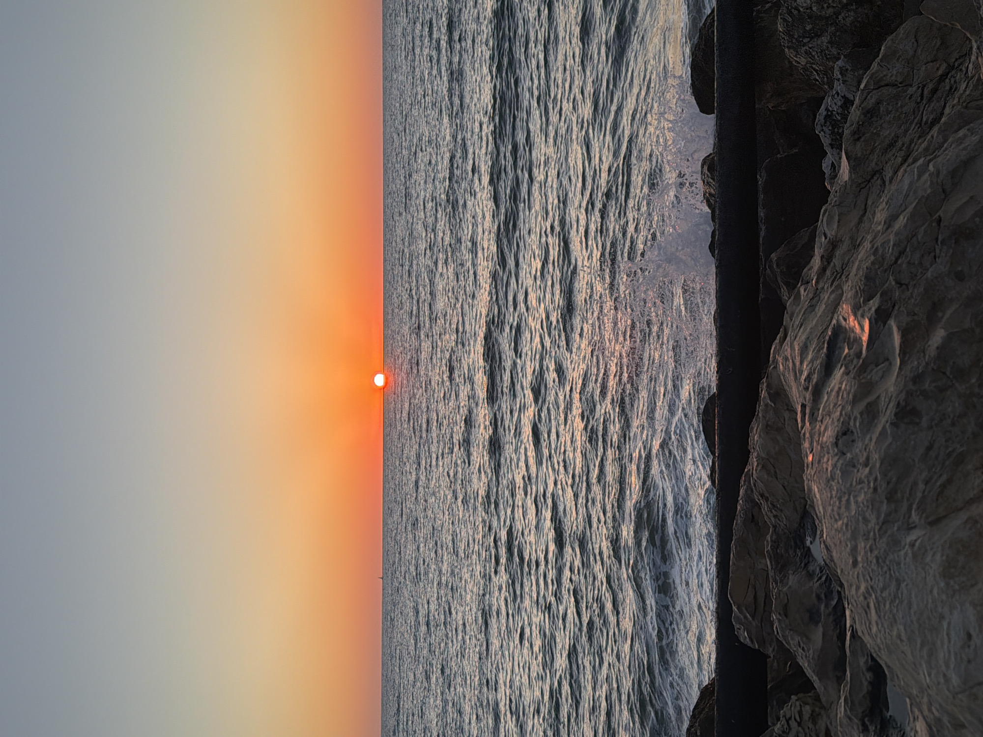 The image displays a serene sunset over the ocean, viewed from a rocky coastline or jetty. The sun, a vibrant orange orb, is positioned low on the horizon, casting a warm, luminous glow across the scene. The sky transitions from rich orange and yellow hues near the sun to softer, lighter blues and greys higher up.
The vast expanse of the sea is characterized by numerous small waves and ripples, which catch and reflect the ambient light, creating shimmering patterns on the water's surface. On the right side of the frame, dark, rugged rock formations or a sturdy sea wall provide a stark, textured contrast against the sparkling water and expansive sky. A tiny, almost imperceptible dark silhouette, possibly a distant boat or buoy, can be seen on the horizon line in the distance.
The time of day is clearly sunset, indicated by the low position of the sun and the warm, fading light. The weather appears clear and calm, allowing for an unobstructed view of the horizon and smooth water. This setting is located in Tel-Aviv, Israel, overlooking the Mediterranean Sea. The scene evokes a sense of tranquility and natural beauty. There is no visible text in the image.
