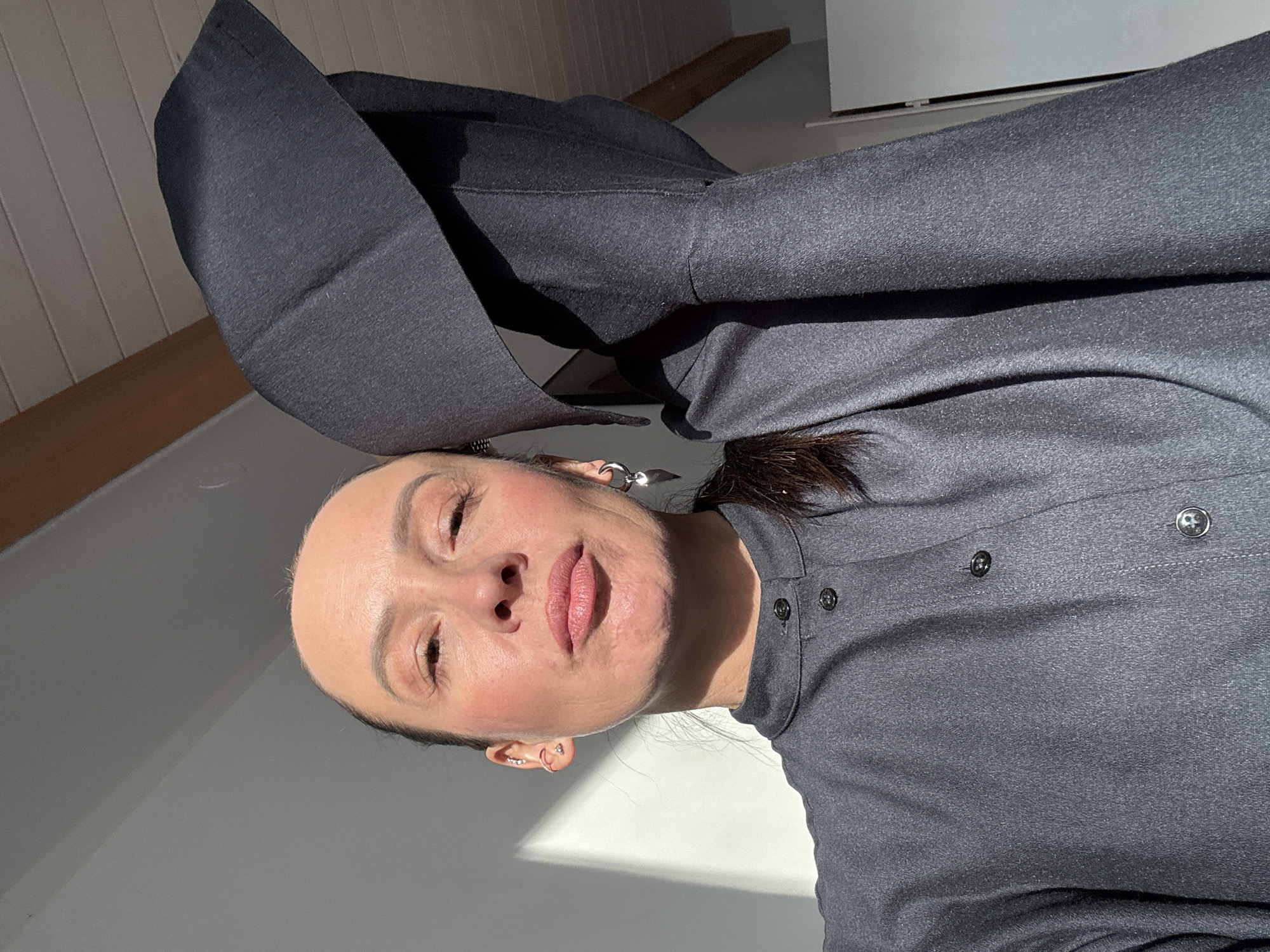 A woman poses for a selfie indoors. She wears a gray shirt with visible buttons. Part of a garment with a large hood is visible behind her. The background includes parts of a ceiling and a wall. It's daytime, evidenced by the lighting. There is no information in the image to confirm its location in Chaikovsky, Russia.