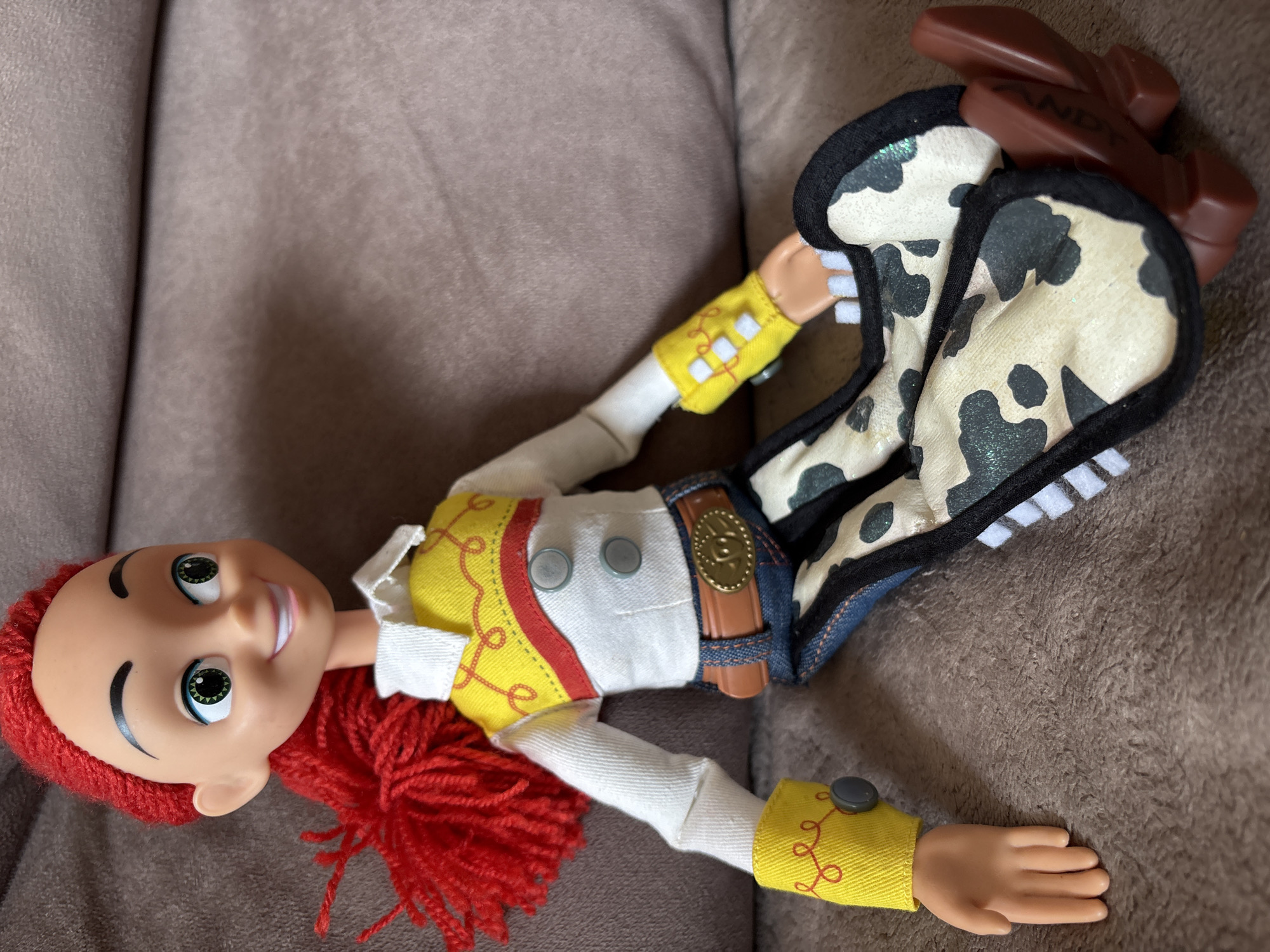 The image shows a close-up of a Jessie doll from Toy Story, dressed in her iconic cowgirl outfit. The doll has bright green eyes, red yarn hair, and a wide smile. She is wearing a white shirt with a yellow and red vest, blue jeans, and cow-print chaps. The doll is sitting on a brown, textured surface, possibly a couch or blanket. The lighting is natural, casting soft shadows. There is no indication of the doll's location in Ветлужский, Russia.
