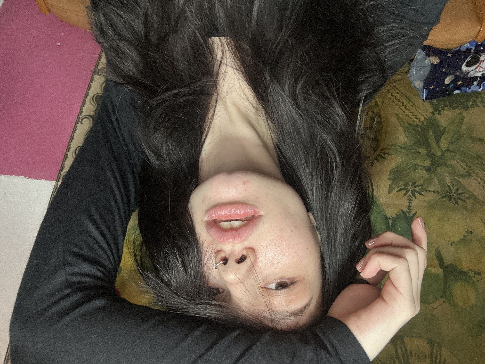 This image presents a close-up, indoor portrait of a female individual. She has long, dark, wavy hair, which partially obscures her face. Her brown eyes are directed towards the viewer, and her lips are slightly parted, revealing some lower teeth. A silver-colored, circular ring is visible in her septum piercing. Her facial skin exhibits minor blemishes. The individual's left arm is raised, with the forearm and hand visible, resting atop her head and hair. Her fingernails are painted with a light-colored, possibly glittery polish. She is clad in a black, long-sleeved garment.
The background on the left side of the frame features a green wall covering or fabric with a repeating stylized leafy or floral pattern. Below this, a fragment of a blue item patterned with white astronauts and space elements is visible. Further below, a brown, textured surface, possibly an armchair or sofa, is present. To the right, a solid pink wall occupies a significant portion of the background, separated from the green patterned area by a narrow, light-colored strip with a curvilinear pattern. The scene is located in Aktanysh, Russia.