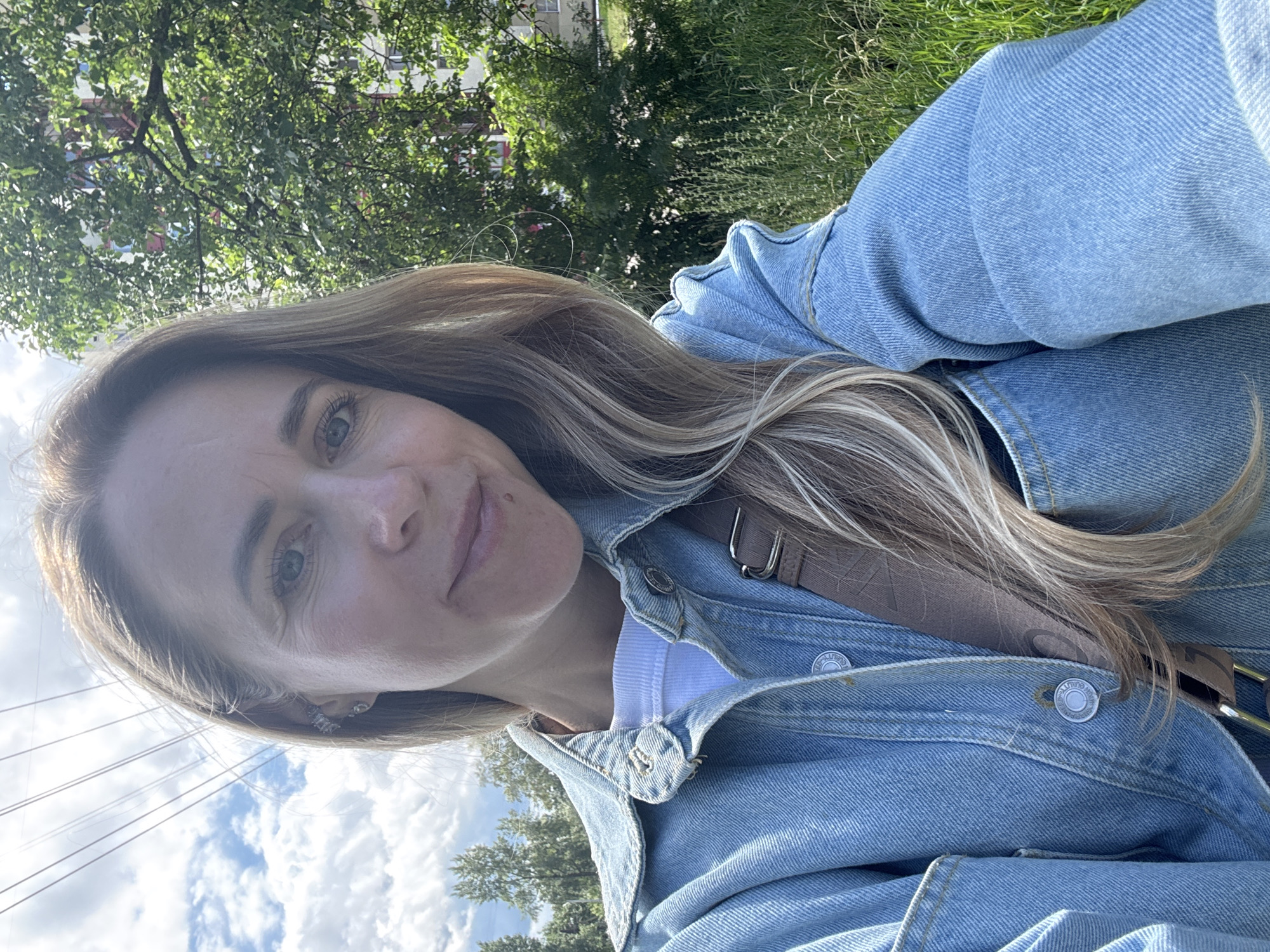 This is a selfie taken by a woman outdoors. She has blonde hair with highlights and is wearing a light blue denim jacket over a white t-shirt. A brown strap from a bag is visible across her shoulder and jacket. The background shows a tree with green leaves, green grass, and a cloudy blue sky with some telephone wires visible. The lighting suggests it is daytime, likely sunny. The woman's expression is neutral. The image is oriented sideways.