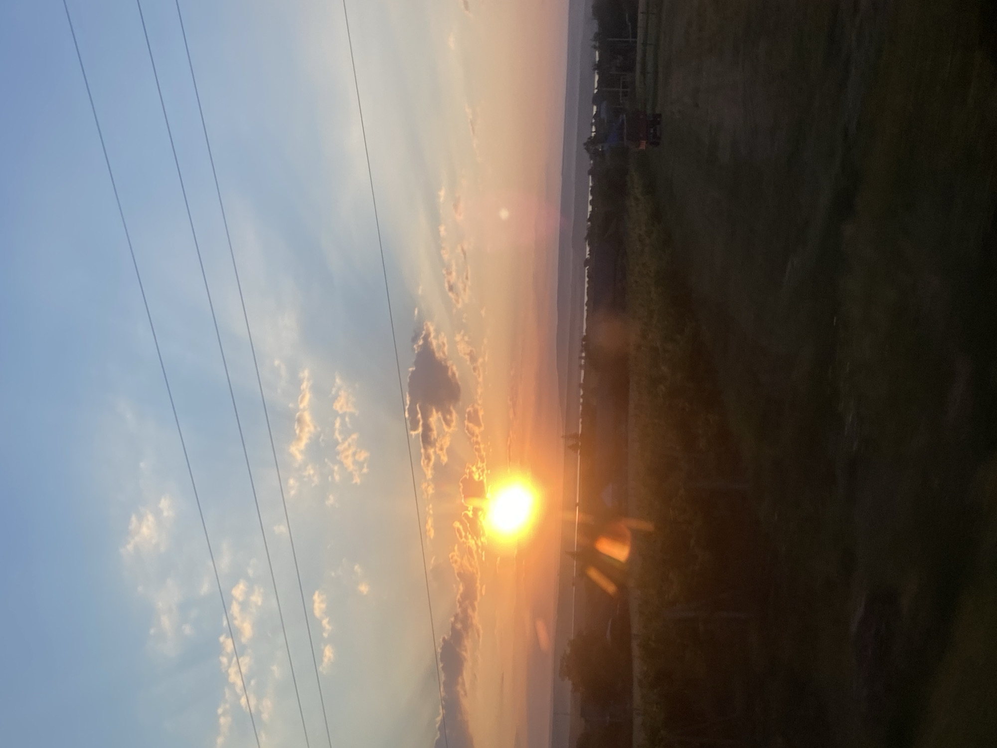 The image captures a sunset over the rural landscape of Balabanu, Moldova. The sun, a bright orb, is low in the sky, casting warm orange and yellow hues across the clouds and the horizon. Rays of sunlight fan out from the sun, illuminating the sky and creating lens flare effects. In the foreground, there is a field of crops, possibly a vineyard, with some vertical stakes visible. Further in the distance, there are scattered houses and trees, suggesting a rural settlement. A lone vehicle with a trailer is visible in the field on the right side of the image. The sky transitions from a clear blue at the top to a warm gradient of orange and pink near the horizon. Several power lines are stretched across the upper portion of the image. The overall scene depicts a serene, rural evening in Moldova.