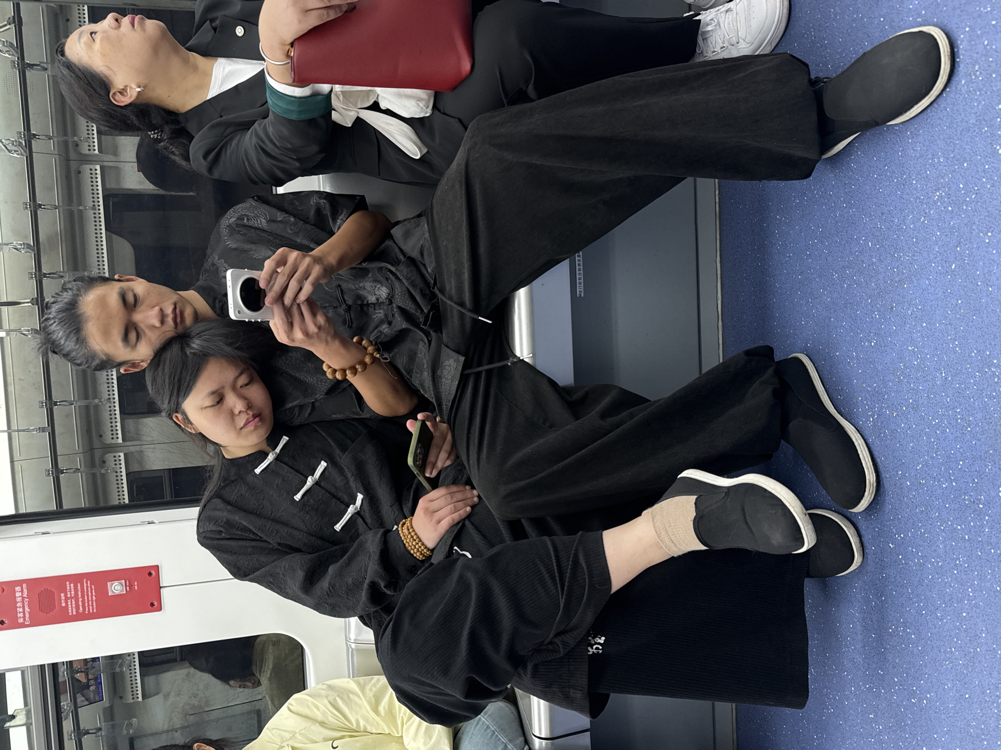 In Yuzhong District, China, a crowded subway car features three individuals seated. A woman in a black business suit with white sneakers leans back with her eyes closed, holding a red object, possibly a tablet or file. Seated next to her, a man with a topknot and wearing a black traditional outfit is using a white smartphone. His head rests on the head of a woman seated beside him, who is also clad in a black traditional outfit. She is holding a green smartphone. The floor of the subway is blue with white specks, and a red sign with white lettering is visible on the wall. The overall atmosphere suggests a scene of people commuting during travel.
