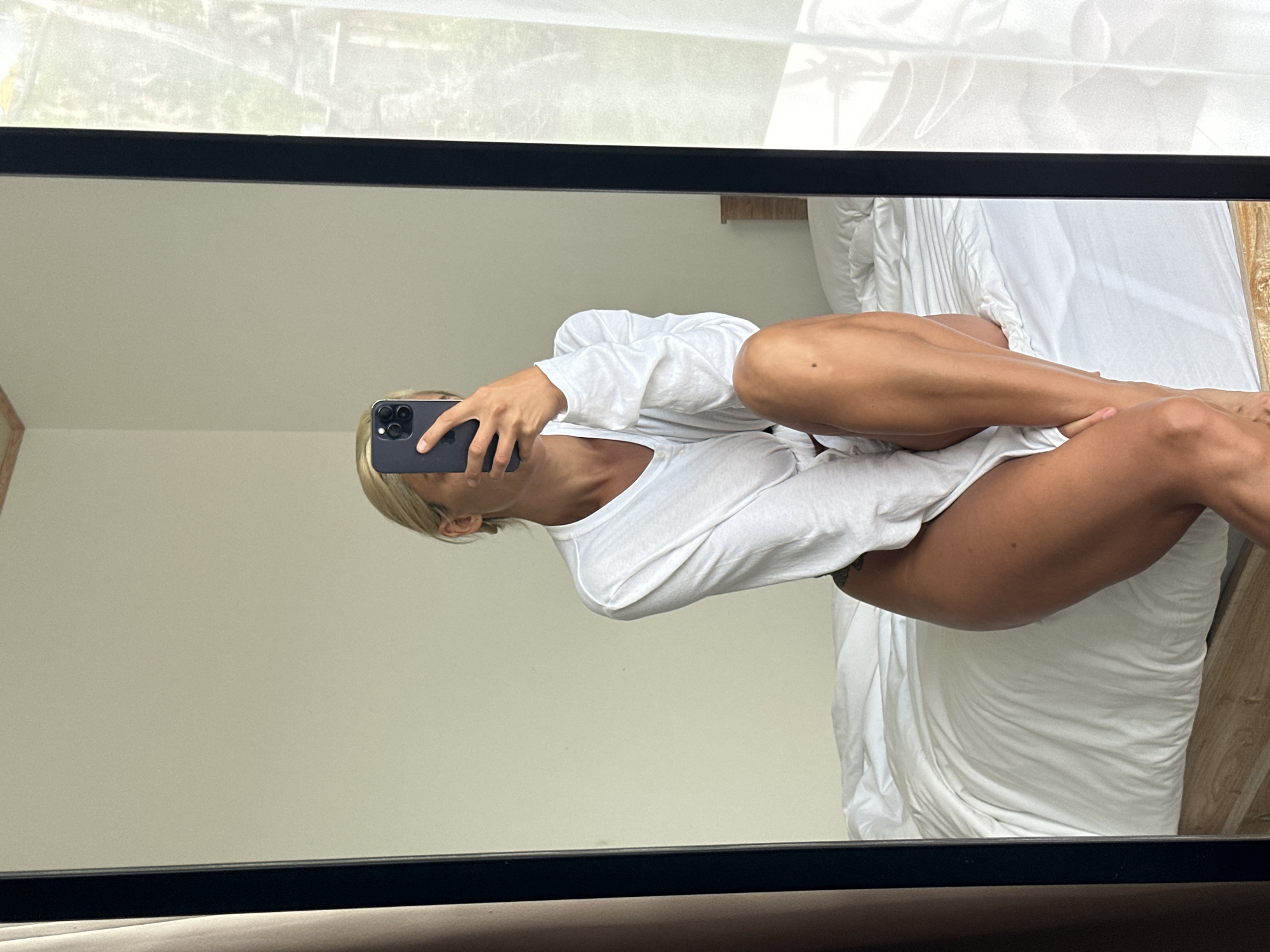 A woman with blonde hair is sitting on a bed, taking a mirror selfie. She is wearing a white long-sleeved shirt. The bed has white linens and a wooden headboard. The room appears to be a bedroom, possibly in a hotel or resort, with light-colored walls and sheer curtains visible through the mirror's reflection. The overall setting suggests an indoor residential or hospitality environment, likely in a warm climate given the sheer curtains and ambient light.