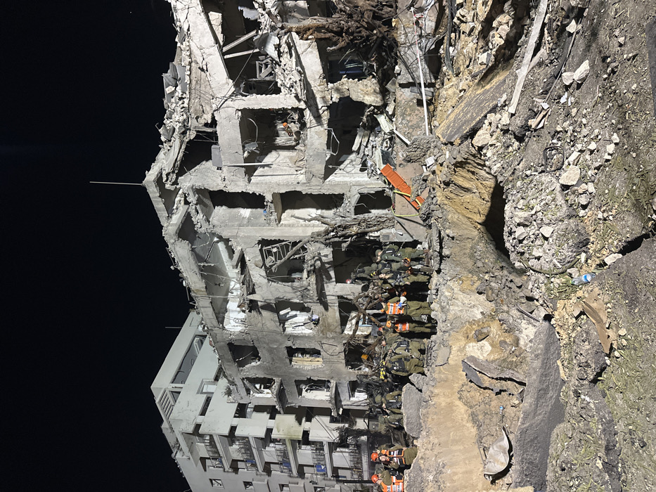 The image depicts a nighttime scene in the city of Tel-Aviv, Israel, dominated by a severely damaged multi-story building. The structure, likely residential, exhibits extensive partial collapse, with its facade and internal sections reduced to exposed concrete, rebar, and rubble. To the left, an adjacent, intact multi-story apartment building with balconies is visible, illuminated against the dark sky.
In the foreground, a substantial amount of debris and excavated earth fills the ground, forming a large crater-like depression or a mound of rubble. A group of approximately 20-30 individuals is gathered near this debris, directly in front of the collapsed building. Many are wearing olive green or camouflage military-style uniforms, often augmented with bright orange or yellow high-visibility vests, suggesting they are search and rescue personnel, possibly from military or civilian emergency services. Some individuals appear to be communicating or observing the scene. An orange stretcher or backboard is visible leaning against the rubble on the right side of the group. Exposed tree roots are also visible amidst the debris, indicating significant ground disturbance. The scene is illuminated by artificial light sources, casting shadows and highlighting the destruction. A discarded blue plastic water bottle lies on the ground in the immediate foreground.