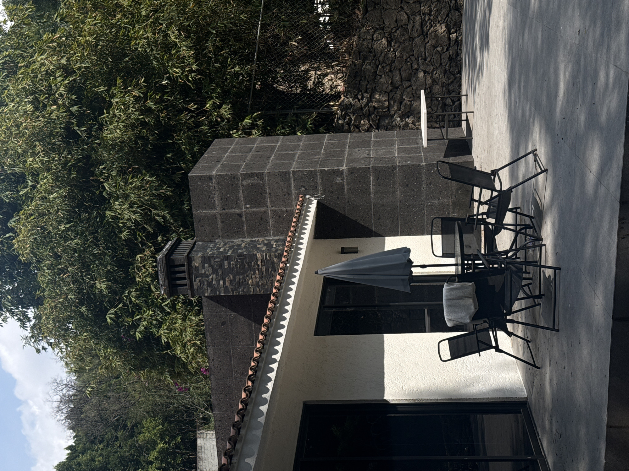 The image shows an outdoor patio scene in San Bernardino Tlaxcalancingo, Mexico. The patio is constructed of light gray concrete and features a dark gray outdoor dining set consisting of a table and several chairs, some with white towels draped on them. An umbrella is partially open over the table. The building adjacent to the patio has a white exterior wall with windows. A brick chimney rises from the roof. The patio is bordered by a dark gray stone wall and chain link fence, behind which is a dense thicket of trees and foliage.