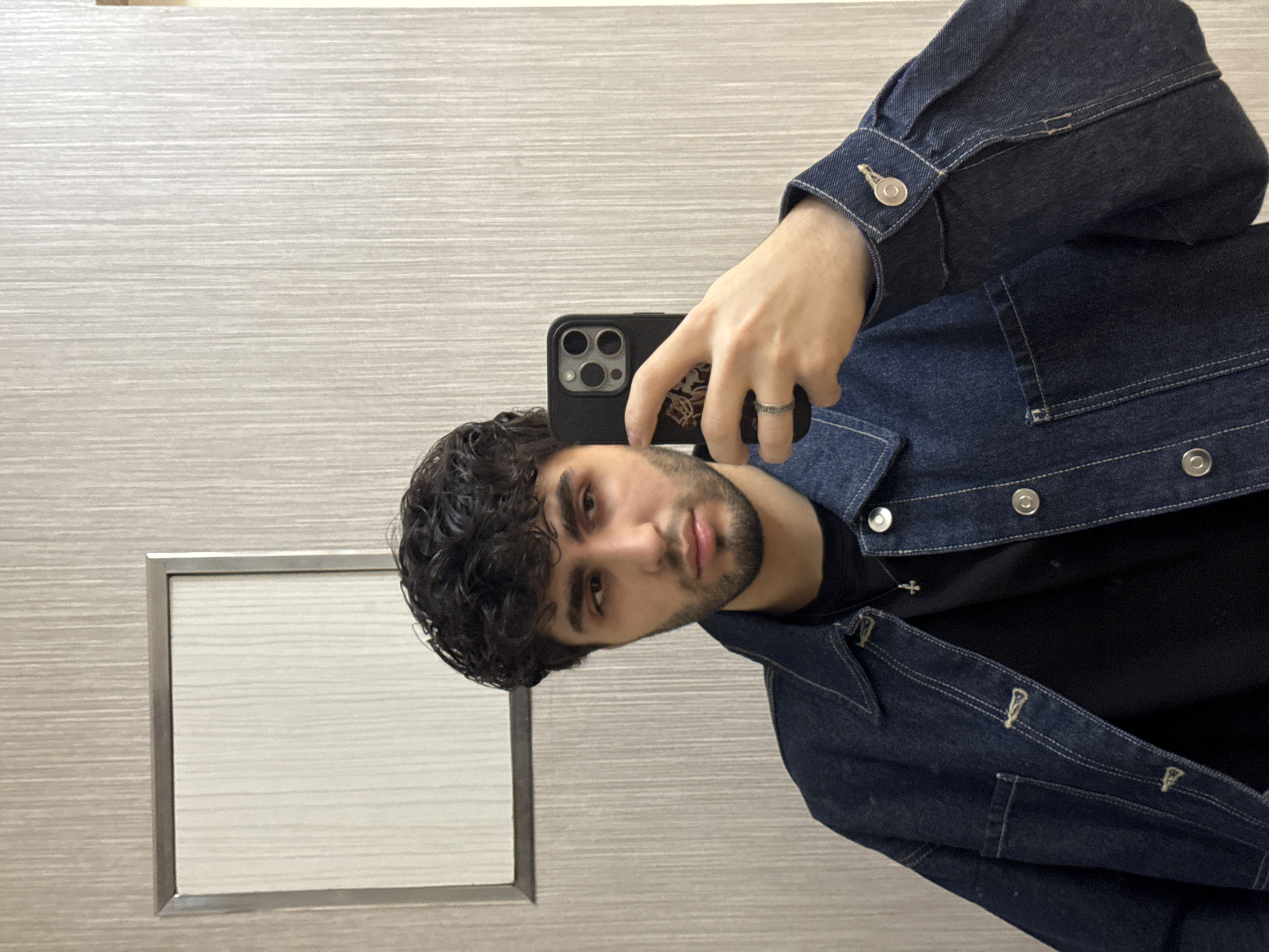 A young man with dark curly hair and a neatly trimmed beard poses for a selfie in what appears to be a restroom. He is wearing a black t-shirt and a dark blue denim jacket. The background consists of a light grey wall with horizontal striations and a framed rectangular inset, possibly a mirror or another panel. The man is holding a smartphone with a dark case to take the photograph, and there is a ring on one of his fingers. The location in Yerevan, Armenia, is not directly discernible from the image.