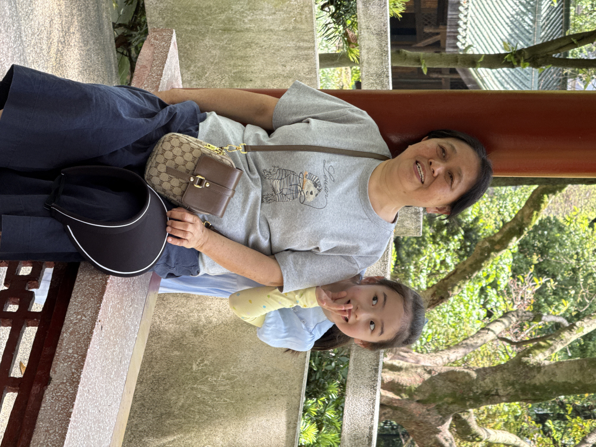 In Yuzhong District, China, an adult woman and a young child are sitting next to each other on a stone bench within a covered structure. The woman has short, dark hair and is wearing a gray t-shirt and dark pants. A patterned brown purse with a brown leather accent is resting on her lap. A black visor is beside the purse. The child, wearing a blue top with yellow sleeves, is making a hand gesture near her mouth. In the background, trees are visible.
