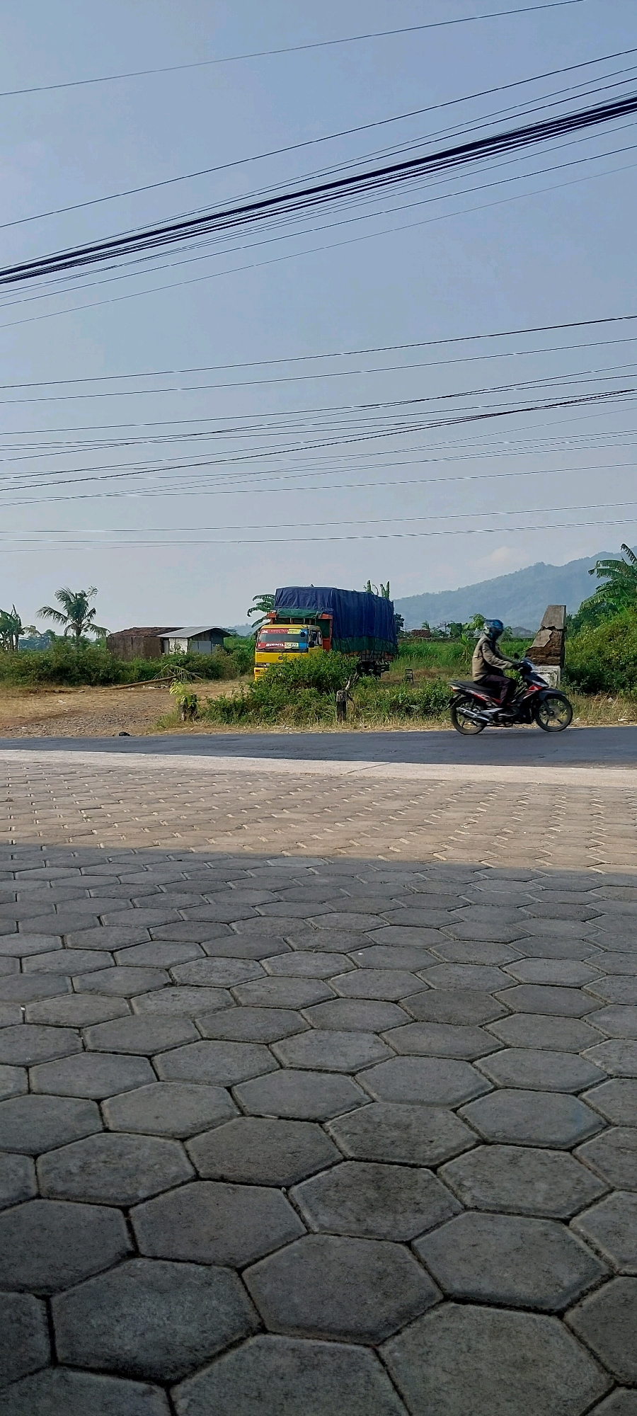 The image depicts a street scene in Keling, Indonesia. The foreground features hexagonal paving stones, indicating a pedestrian area or walkway. In the middle ground, a paved road runs horizontally. To the right of the road, a person is riding a motorcycle, wearing a helmet and a jacket. Further into the background, a large truck with a blue tarp covering its load is parked or stopped on the side of the road. Several small buildings and lush green vegetation, including palm trees, are visible behind the truck. The background is dominated by a clear sky and rolling hills or mountains, suggesting a semi-rural or suburban setting. Power lines are strung across the top portion of the image. The lighting indicates it is daytime, with sunlight casting shadows.