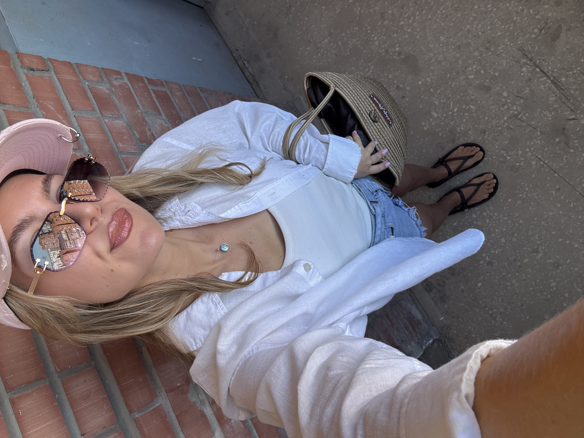 A blonde woman, seen from a high angle, poses for a selfie in front of a brick wall. She wears a pink baseball cap, large sunglasses reflecting buildings, a white linen shirt unbuttoned over a white tank top, denim shorts, and black sandals. She holds a woven straw bag. She stands on a paved surface.