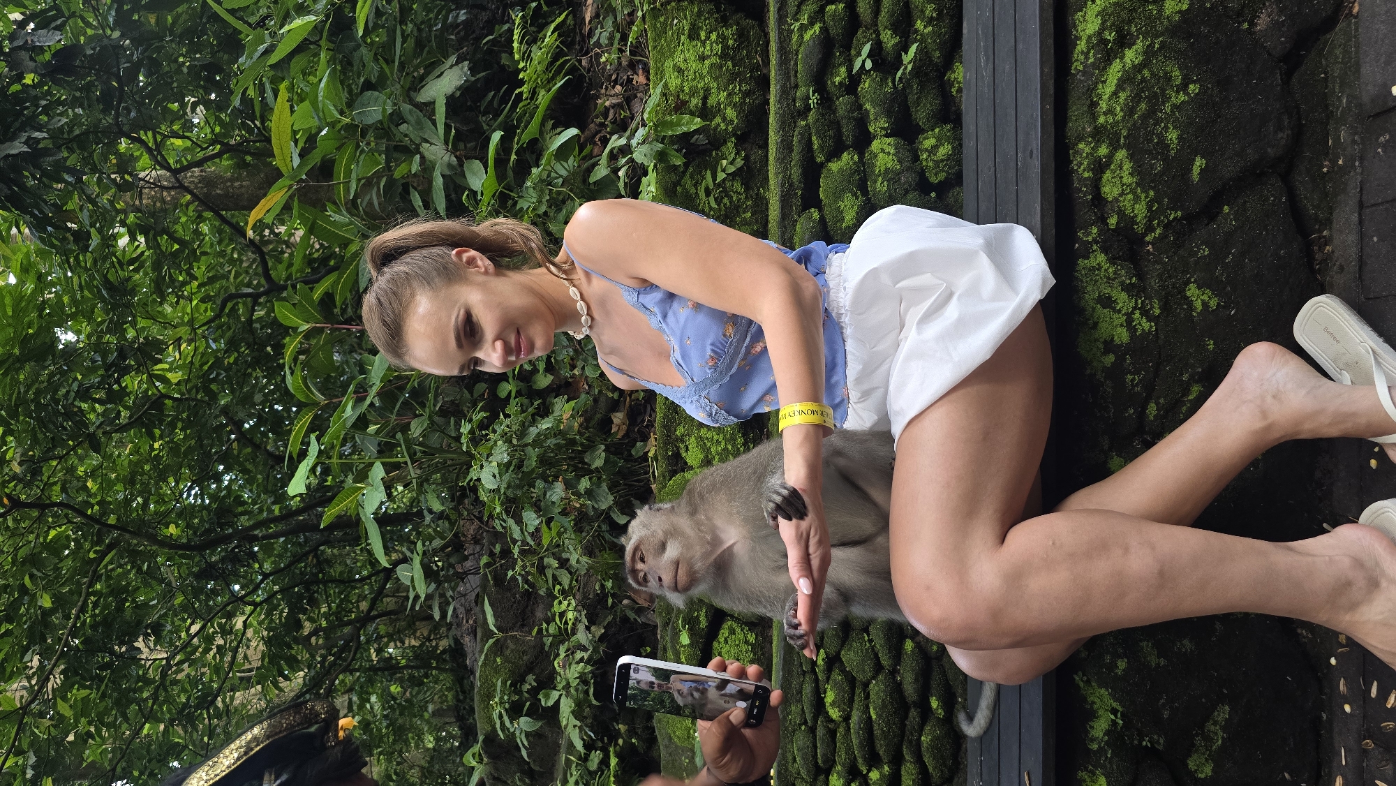 A fair-skinned woman with her light-brown hair pulled back in a ponytail kneels while interacting with a monkey at a park in the Ubud District, Indonesia. She wears a light blue floral top with lace trim, white shorts, and a pearl necklace. A yellow wristband is visible on her right wrist. A monkey is seated next to her and appears to be holding her right hand. A person, partially visible, is taking a picture of the interaction with a smartphone. The environment consists of moss-covered rocks, various plants, and trees.