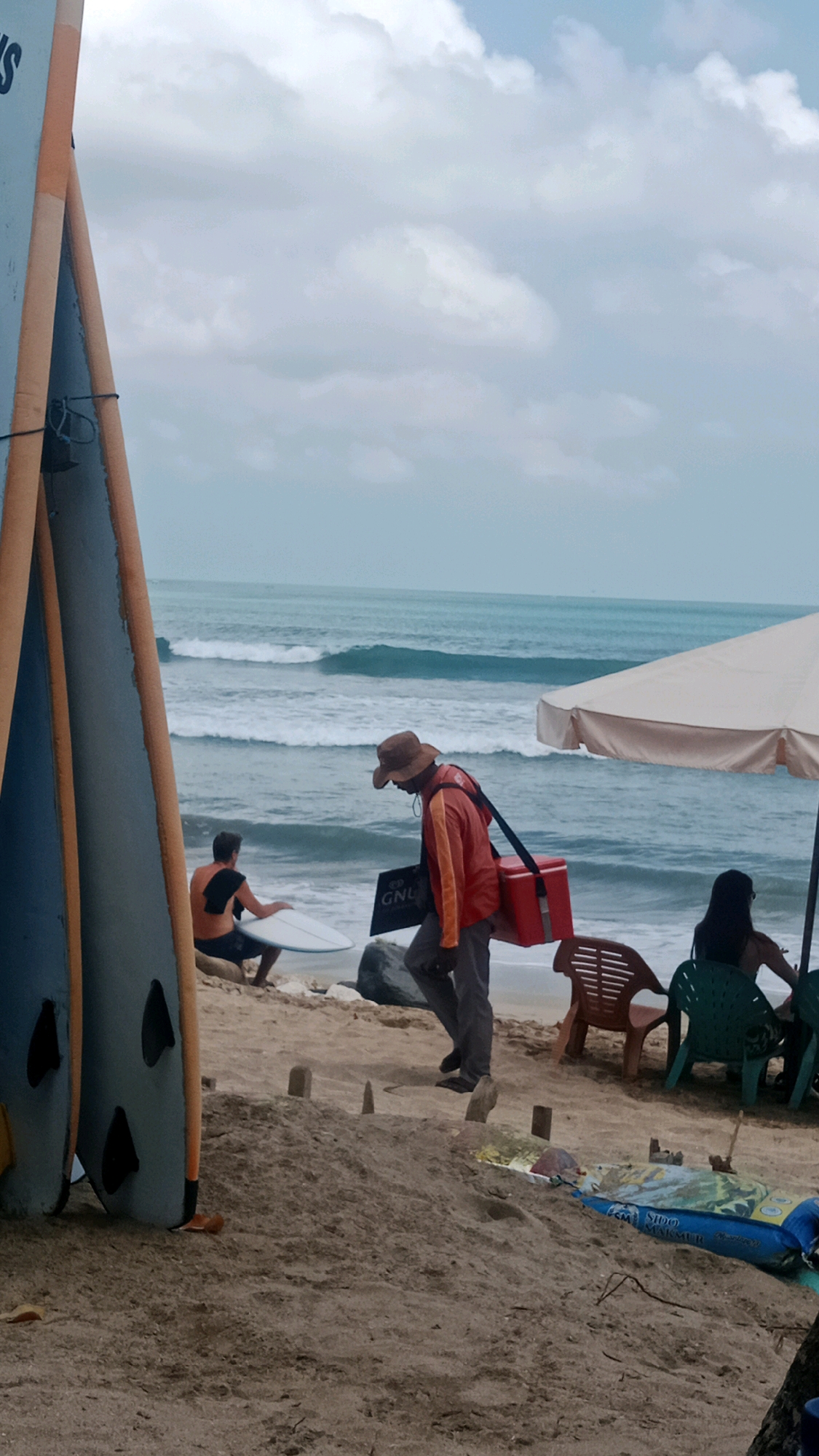 A group of people are on the beach in Kuta, Indonesia, near the ocean. The sky is cloudy, and there are waves in the ocean. In the foreground, there are several surfboards leaning against each other. A man wearing a hat and an orange jacket is walking with a red cooler bag. A person in the middle ground is sitting on the sand with a surfboard. In the background, there are beach chairs and an umbrella. The sand is light brown.
