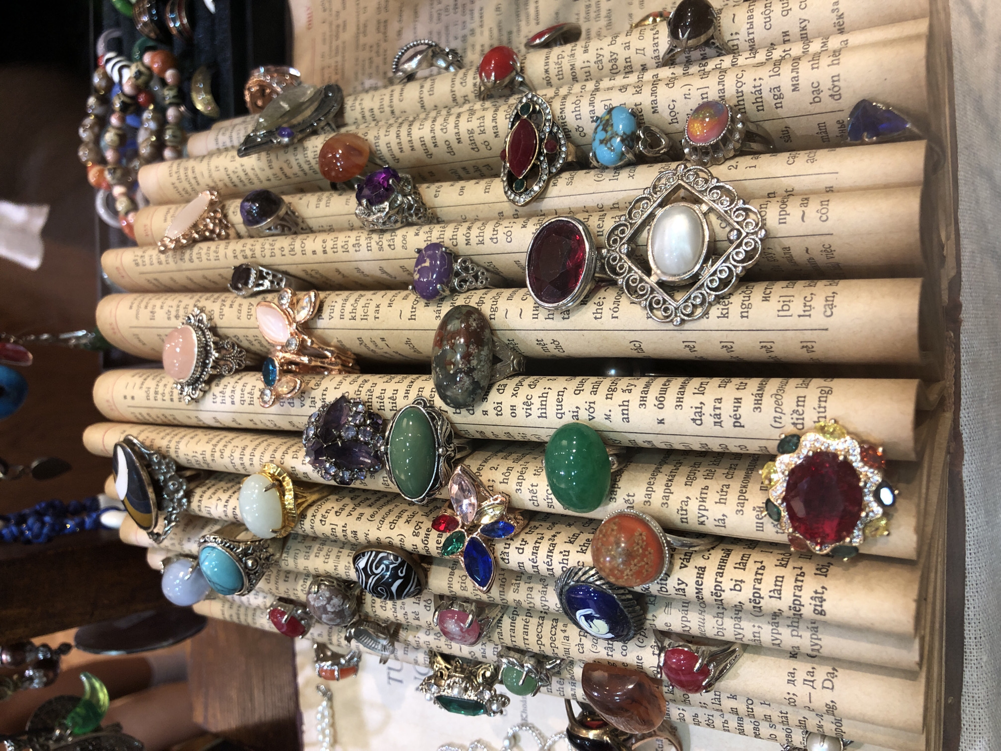 The image shows a close-up, overhead view of a collection of rings displayed on stacks of old books. The rings vary in design, with many featuring colorful gemstones such as amethyst, turquoise, and red stones. Some rings have intricate metalwork, while others have simpler, bolder settings. The books, which appear to be vintage, have pages filled with text in various languages, contributing to a cluttered yet artistic presentation. The overall scene evokes a sense of a vintage jewelry stall or a personal collection. There are no people visible in the image. The lighting suggests an indoor setting, possibly a shop or a display case. The text on the books is predominantly in Vietnamese and possibly Cyrillic, indicating a diverse source or a collection from various regions. The focus is solely on the rings and the aged books, creating an atmosphere of historical charm and handcrafted beauty.