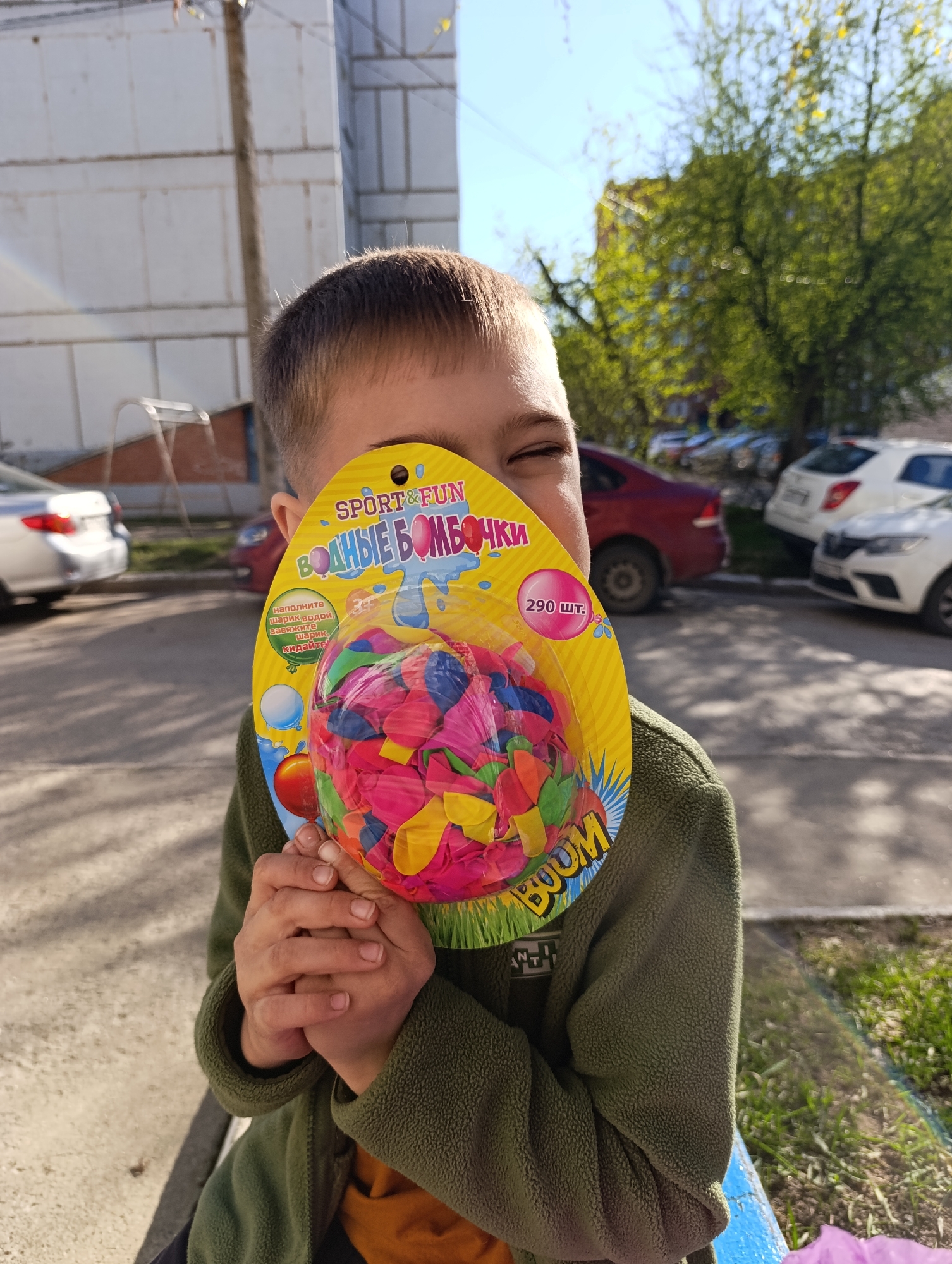 A young boy is holding a package of water balloons in front of his face, obscuring most of it except for one eye. He is wearing a green fleece jacket. The package of water balloons is yellow and labeled in Russian, "ВОДНЫЕ БОМБОЧКИ" (Water Bombs), with "SPORT&FUN" at the top and "BOOM" at the bottom. The package contains an assortment of colorful, uninflated balloons. The boy's hands are clasped together, holding the package firmly. He is located outdoors, seated on a blue painted railing, with cars parked in the background. The image appears to have been taken in the city of Chaikovsky, Russia.