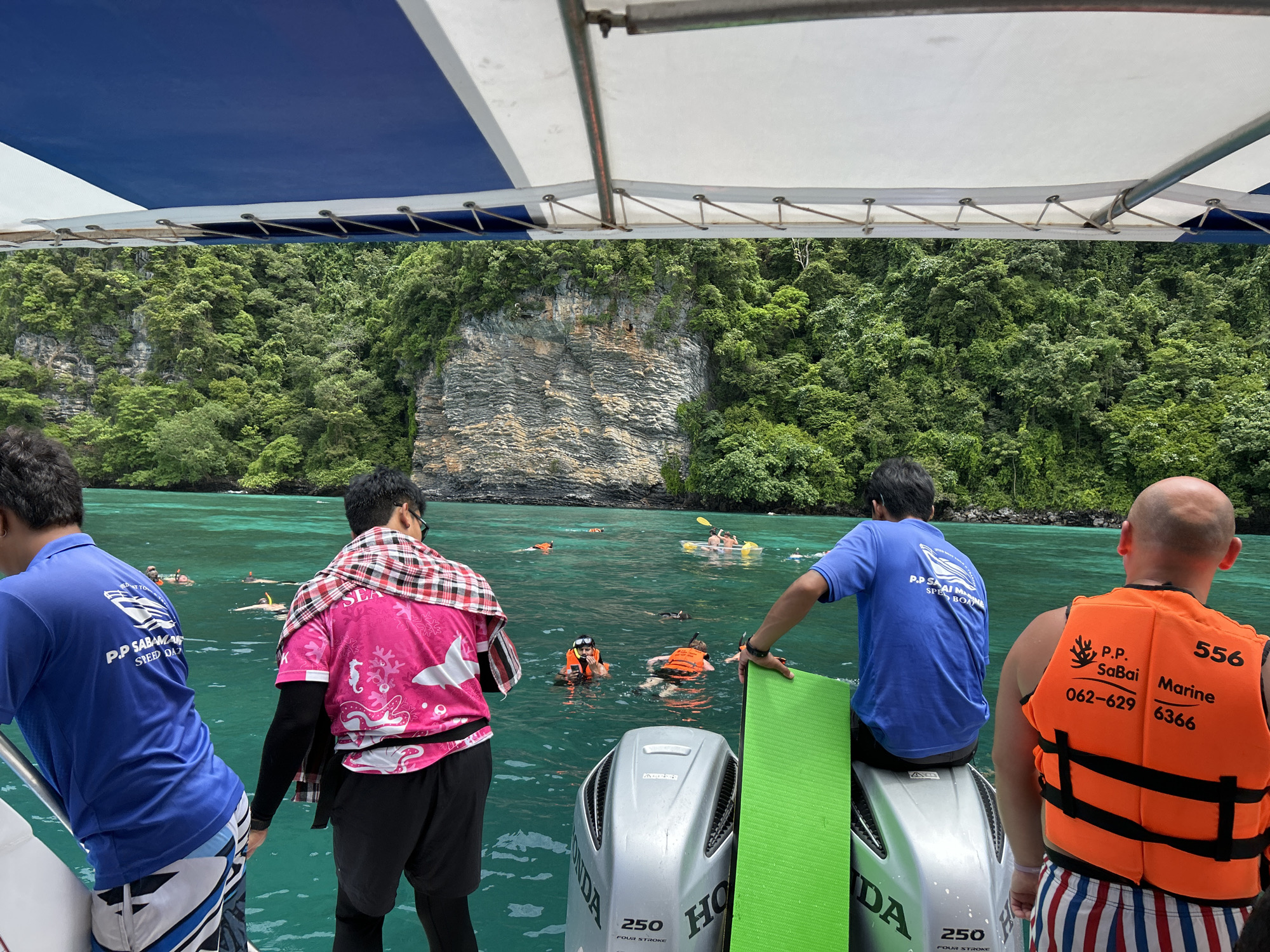 A rear view shows four men on a boat in the turquoise waters off Ao Nang, Thailand. The boat has a blue and white canopy overhead. Three men are visible from behind, wearing casual attire. One man in a blue t-shirt stands to the left, next to a boat engine. Another man in a pink t-shirt with a checkered scarf draped over his shoulders stands in the center. A third man with a bald head wears an orange life vest and stands on the right. In the background, several people are swimming and snorkeling in the clear water near a lush, green cliff face. The scene suggests a day trip or tour in a tropical marine environment.