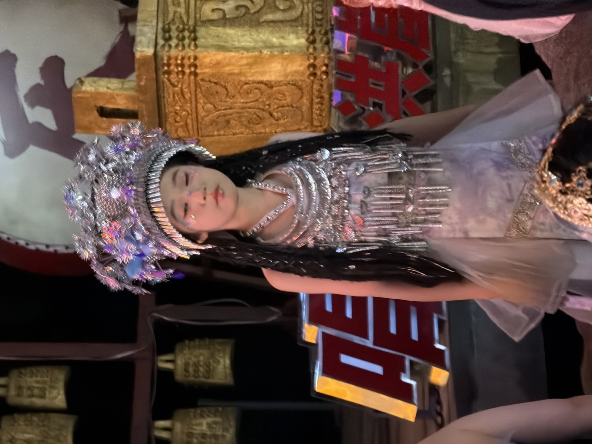 A young girl, likely of East Asian descent, is standing center frame wearing a highly ornate, silver headdress adorned with blue feathers and shimmering accents. She has dark braided hair that extends down her back and is wearing a layered, silver and lavender costume with intricate beadwork and dangling embellishments. Glittery makeup is applied to her face, with accents on her cheeks and eyelids. She appears to be on a stage, with a large, blurred red and white sign behind her that appears to be in Chinese characters. To the left and right of the sign are large, golden, carved structures. In the background, several large, golden bells are visible hanging from a dark structure. The lighting suggests an indoor performance or event. The backdrop elements and signage indicate a potential cultural performance or festival setting.