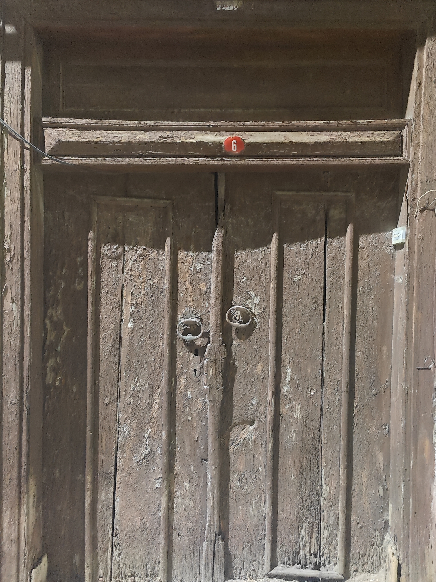 A close-up, low-angle shot captures a weathered, brown wooden double door in Osmangazi, Turkey. The door is divided vertically into two panels, each featuring raised vertical stiles. The wood exhibits significant wear and peeling paint, revealing a lighter wood underneath in many areas. Two ornate, circular metal door knockers are centrally placed on each door panel, with a visible keyhole below them. Above the door, a horizontal wooden ledge has a small, oval red sign with the white number "6" adhered to it. The top of the door is recessed, with a darker wooden frame. A thin black wire runs diagonally from the upper left corner of the image across the door's frame. A light-colored electrical outlet is mounted on the right side of the door frame. The lighting suggests an outdoor setting, with strong shadows falling across the door's surface.