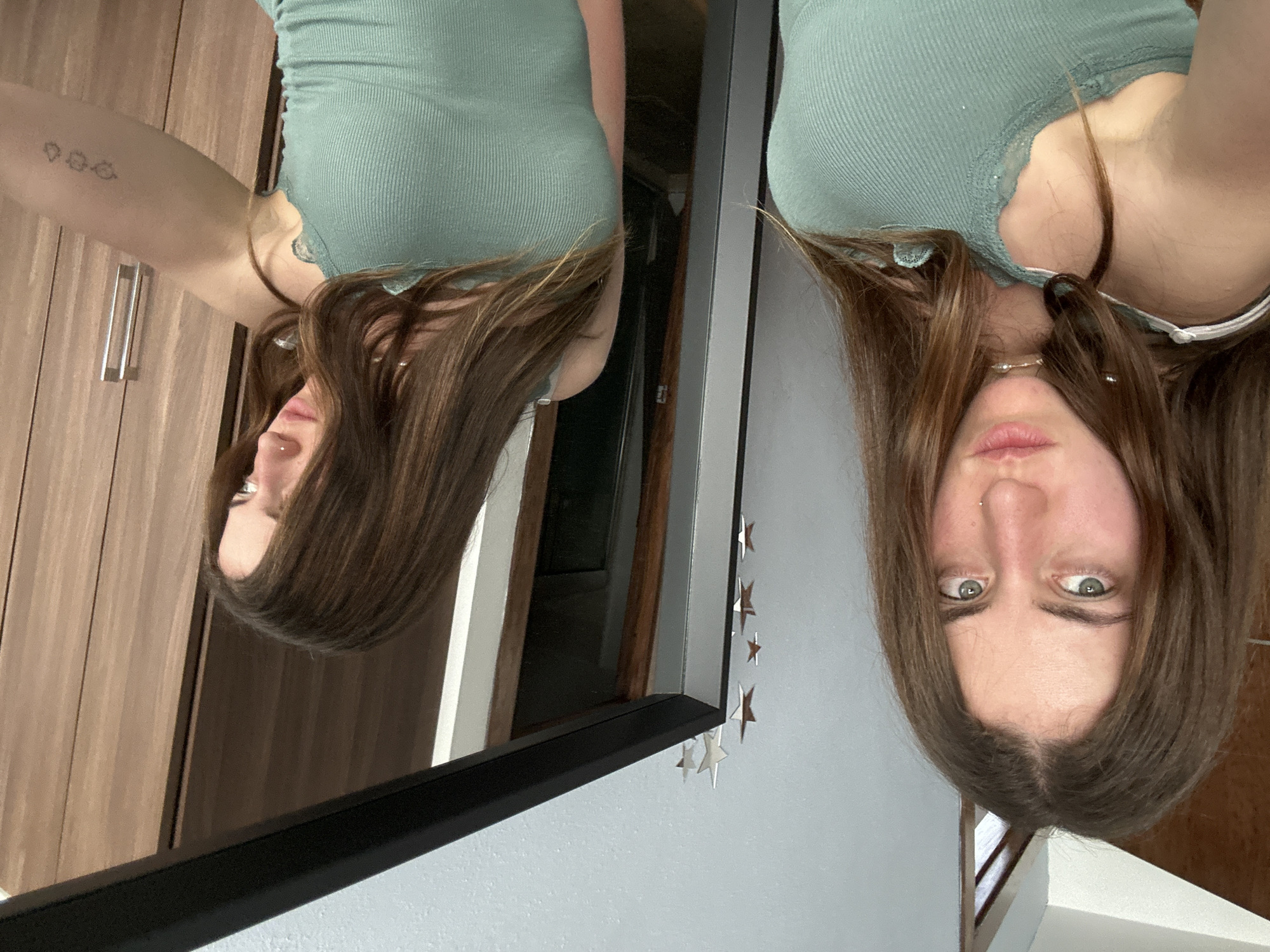 A woman is reflected in a mirror. The woman is wearing a green tank top with lace trim. She has long brown hair and green eyes. She has a nose ring. Her right arm has a tattoo. The background includes a wooden cabinet and a white wall decorated with silver star stickers. The photograph was likely taken indoors. There are no indications of the location being in Huixquilucan de Degollado, Mexico.