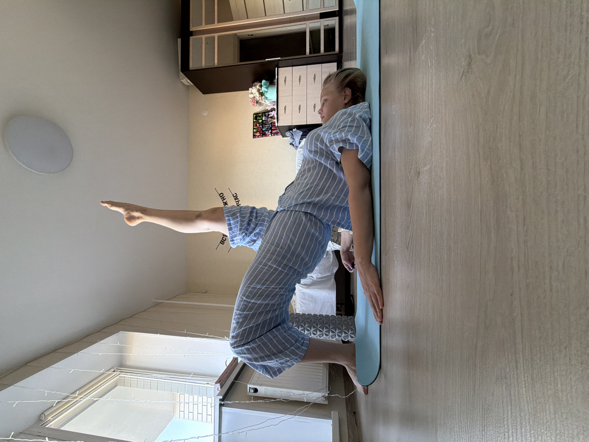 A woman in blue and white striped pajamas is performing a yoga pose on a light blue yoga mat in what appears to be an apartment. She is kneeling with her back arched and one leg extended upwards towards the ceiling. The apartment has light wood-look flooring and light beige walls. A white circular ceiling light is visible, along with a window that has what appears to be a drying rack with hanging items. A dark wood cabinet is visible on the wall with lighter drawers. Decorative items are on the wall including what appears to be a floral arrangement, photographs and post cards. Based on the writing on the woman's pajamas and the overall setting, this location is likely in Izhevsk, Russia.