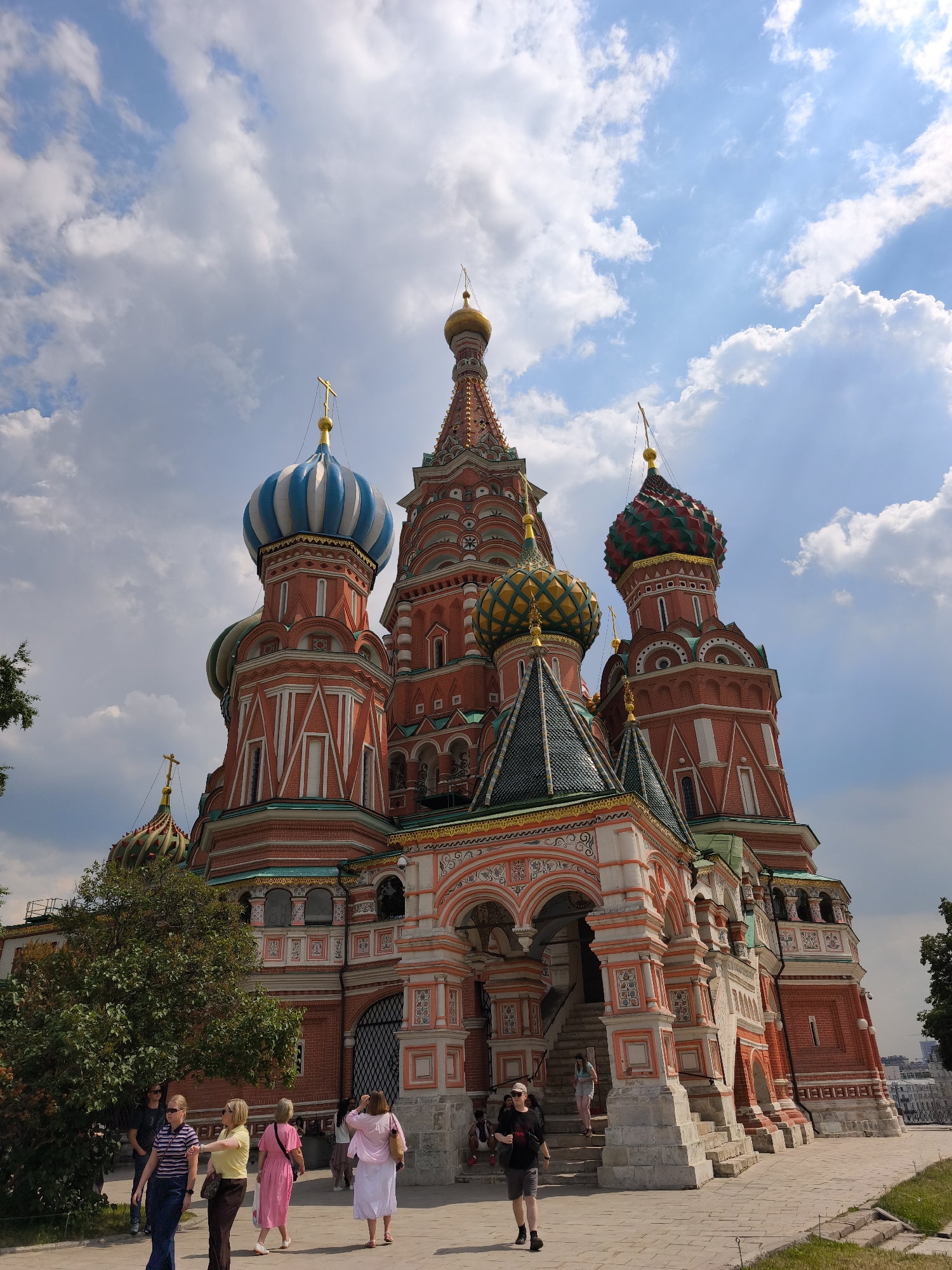 The image captures a low-angle, full-shot of Saint Basil's Cathedral in Moscow, Russia, under a partly cloudy sky. The cathedral's iconic onion domes, painted in red, white, blue, green, and gold, are the primary focal point. The intricate architecture, characterized by vibrant colors and detailed ornamentation, is prominent. Several tourists are visible walking on the paved plaza in front of the cathedral. Some are gathered near the entrance, while others are strolling across the area. A large green tree is situated to the left of the frame, partially obscuring a section of the cathedral. The lighting suggests it is daytime, with sunlight casting shadows and illuminating the scene.
