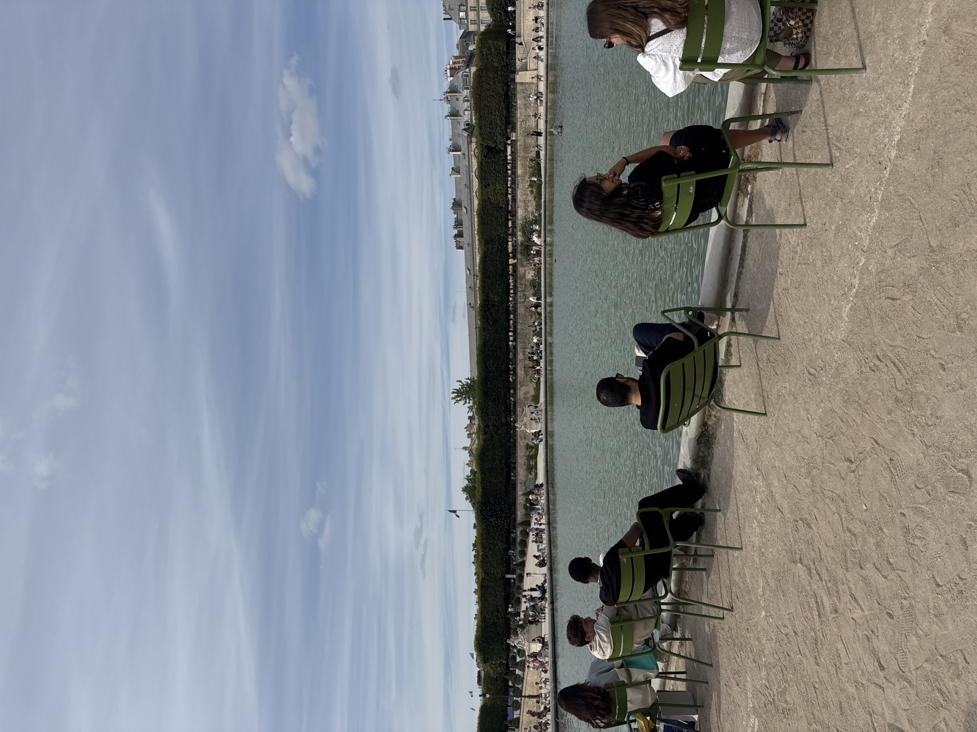 In the Jardin des Tuileries in Paris, France, several people are seated on green metal chairs facing a large pond on a sunny day. In the foreground, from left to right, a woman with dark hair is wearing a gray top and jeans. Next to her, a young man with curly brown hair is wearing a light-colored hoodie and white pants. To his right, a man with dark skin is wearing a black t-shirt and black pants. Further to the right, a woman with dark hair is wearing a black t-shirt and black shorts. Beside her, a woman with light brown hair is wearing a white lace top and olive green pants. The chairs are arranged along a sandy pathway bordering the pond. In the background, beyond the pond, is a manicured hedge with trees, and behind that, a long building with classical architecture, possibly part of the Louvre Museum complex, is visible under a partly cloudy sky. People are scattered throughout the background, walking and sitting near the pond and the hedge. A French flag is visible flying on a pole in the background. The overall scene depicts a tranquil day in a Parisian public garden.