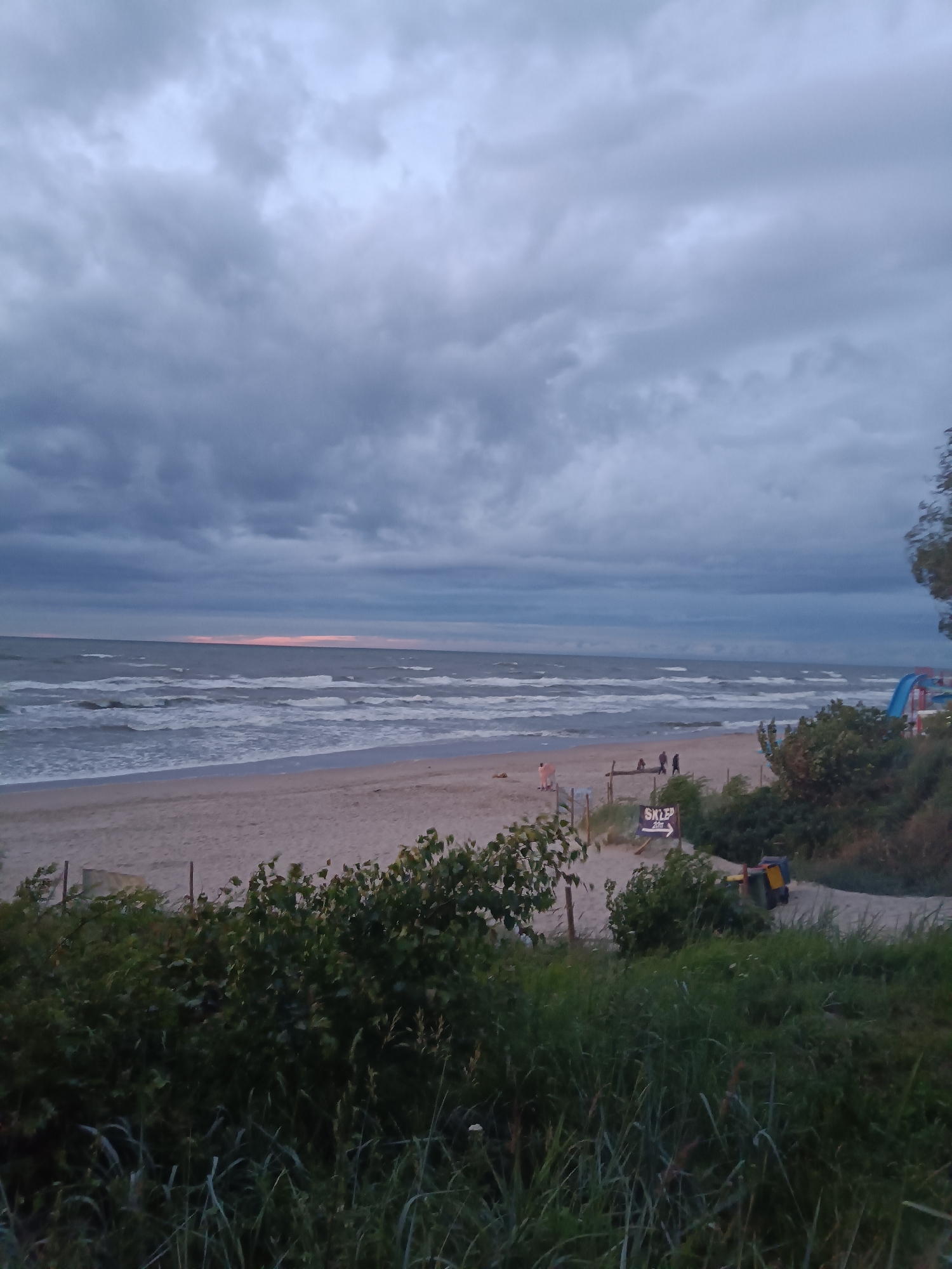 The image depicts a coastal scene at dusk in Stegna, Poland. The sky is overcast with dark, heavy clouds, and the sea is choppy with visible waves rolling towards a sandy beach. A few people are present on the beach, some walking and others standing near structures. A sign indicating "SKLEP 20m" (Shop 20m) is visible, suggesting the presence of amenities. Greenery, including bushes and tall grass, occupies the foreground and parts of the background. A blue and red waterslide structure can be seen on the right side of the image, indicating a recreational area. The overall atmosphere is subdued, characteristic of twilight on a cloudy day.