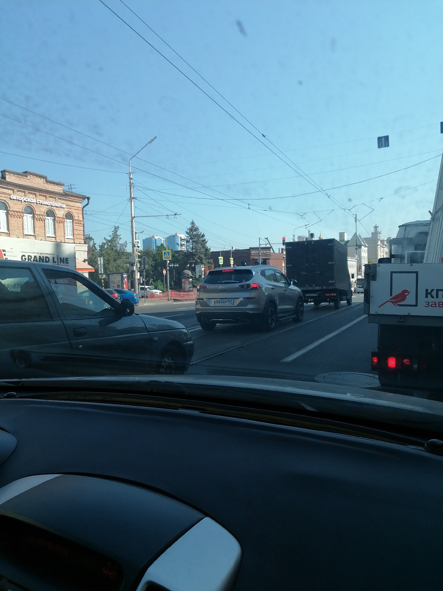 The image depicts a street scene in Rostov-on-Don, Russia, viewed from inside a vehicle. The foreground shows the dashboard and windshield of the car. In the middle ground, a traffic jam is evident with several cars on the road. These include a dark-colored sedan on the left, a silver Hyundai SUV in the middle, and a large truck with a dark box trailer behind it on the right. A white truck with a red bird logo is also visible on the far right. The background features buildings, including a brick building with arched windows and text reading "АВТОРСКАЯ СТОМАТОЛОГИЯ" (Dental Artistry) and "GRAND LINE." Further in the background, modern glass buildings and trees can be seen. The sky is clear and blue with some wires visible overhead, likely from an overhead trolleybus or tram system. Traffic lights and road signs are also present. The overall impression is that of a typical urban traffic scene on a sunny day.