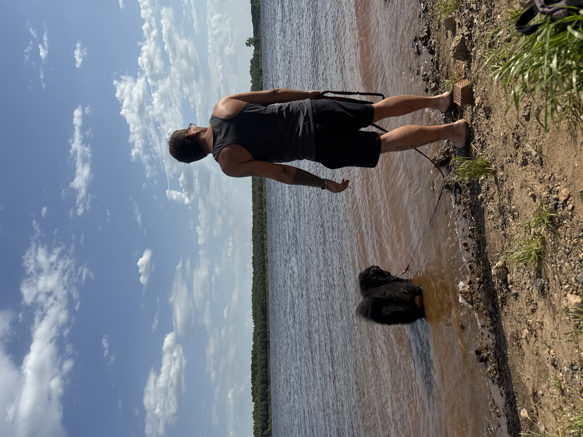 A man and a dog are standing at the edge of a body of water under a partly cloudy sky. The man is fair-skinned, wearing a gray tank top and black shorts. He has short hair and glasses, and a tattoo is visible on his left arm. He is barefoot and holding a black leash. The dog, which appears to be a black Newfoundland or similar breed, is partially submerged in the water. The shoreline is sandy and rocky with some grass. The water is murky brown and extends to the horizon, where a line of trees can be seen. The location is Doksha, Russia.