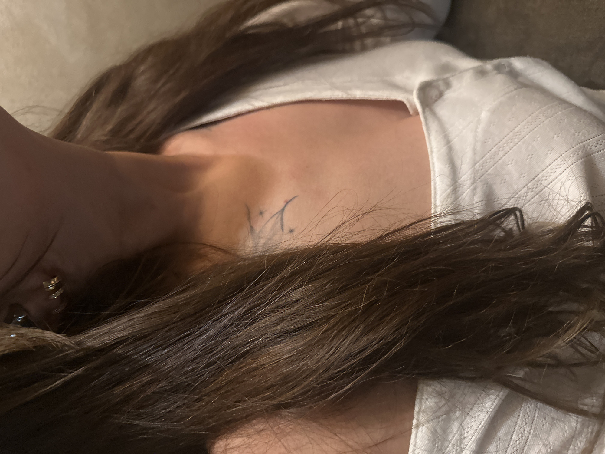 This close-up image, set in Chaikovsky, Russia, captures a person lying down indoors. The central subject is a person, likely female, with long, dark brown hair that spreads across the frame and partially covers their body. They are wearing a light-colored, possibly white, top with a subtle, textured patterned fabric.
A notable detail is a small, dark-colored tattoo located on the person's left clavicle area. The tattoo appears to be a stylized initial or symbol, possibly resembling a 'V' or 'Y', accompanied by delicate decorative elements like small stars or dots. A golden-colored earring is visible on the left side of the frame, near the person's ear.
The activity depicted is the person resting or lying down. Their face is not visible, and the overall impression is one of relaxation. The background behind the person's head is soft and indistinct, suggesting an indoor setting like a bed or couch. The lighting is relatively dim, consistent with an interior space, potentially during the evening or night. No specific weather conditions or other location cues beyond the general indoor setting are discernible from the image.