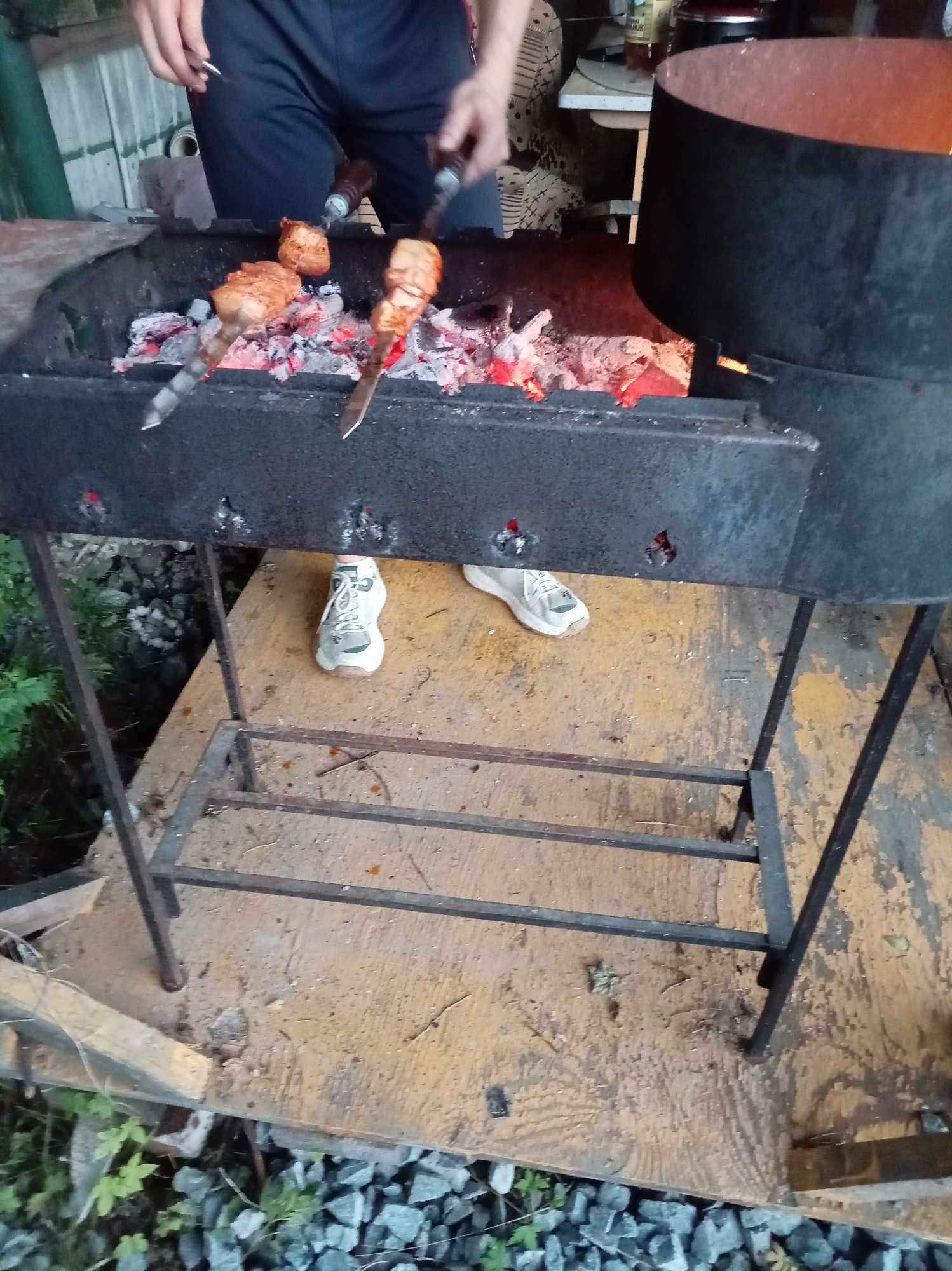 In the city of Artyom, Russia, at an outdoor gathering, a barbecue is in progress. A person, partially visible, is tending to skewers of meat cooking over glowing red coals in a black metal grill. The grill is supported by a black metal frame, and below it is a simple metal shelf. The ground is a wooden platform, covered in scattered debris and stones. In the background, a large black pot, likely for cooking, is situated near the grill. The scene suggests a casual outdoor event, possibly a backyard barbecue.