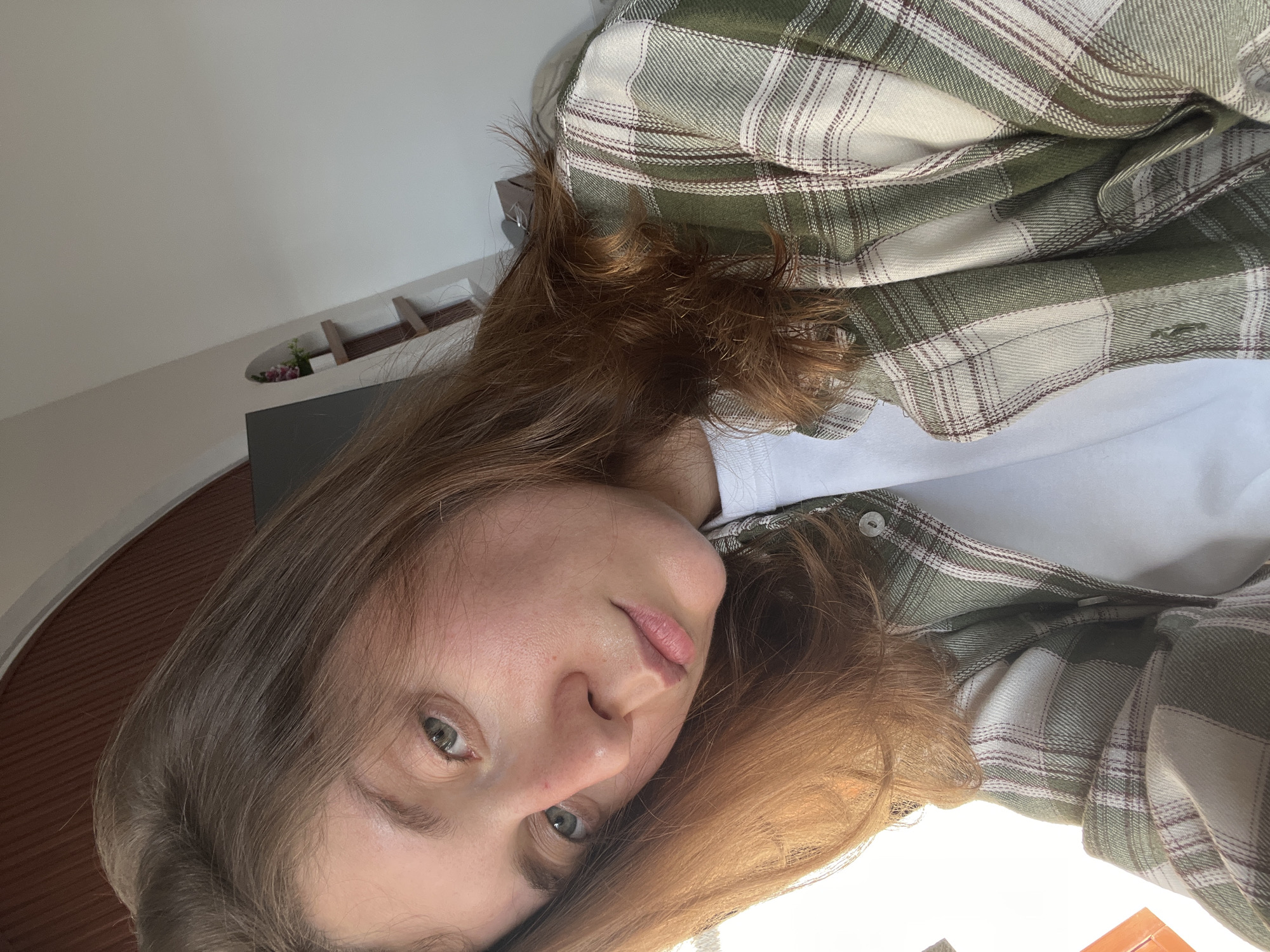 The image shows a person with long, light brown hair, wearing a white shirt under a green and white plaid flannel shirt. The person has light skin and green eyes, and they are looking directly at the camera. The background is out of focus, but it appears to be an indoor setting with light-colored walls and a dark-colored surface. There is a small decorative shelf with a plant on it visible in the background. There is no evidence to suggest this image was taken in North Kuta, Indonesia.