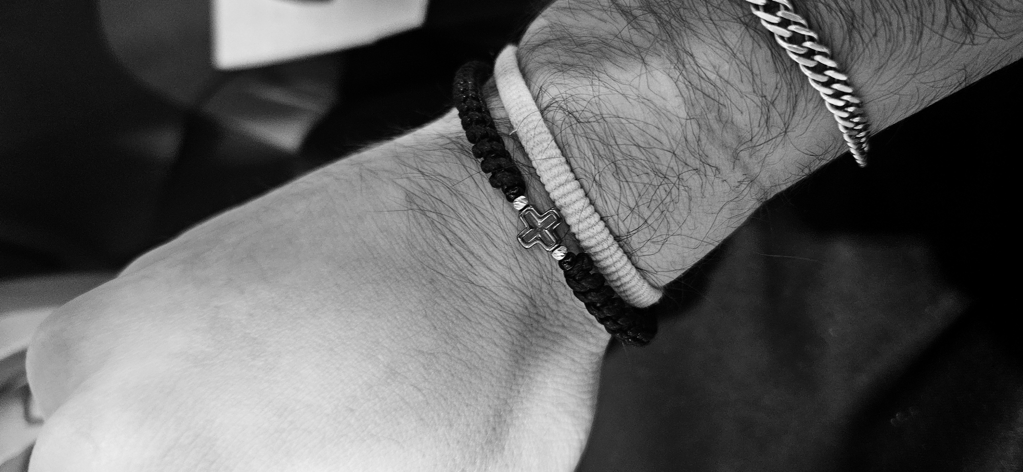 The image depicts a wrist with three bracelets. The bracelets are, from left to right: a black braided bracelet with a silver cross charm, a white rope bracelet, and a silver chain-link bracelet. The wrist is hairy. There is no indication of the location being Plovdiv, Bulgaria.