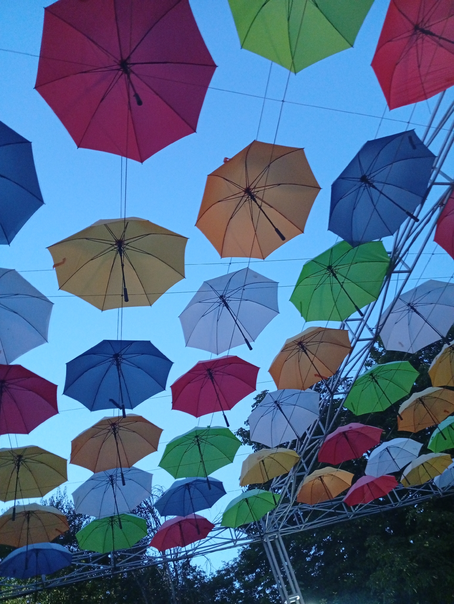 In Pokrov, Ukraine, an outdoor installation features a vibrant canopy of colorful open umbrellas suspended against a clear blue sky. The umbrellas, in shades of red, yellow, green, blue, and white, are arranged at varying heights and angles, creating a visually dynamic effect. Some umbrellas are directly overhead, while others are further away, giving a sense of depth. The scene is framed by the branches of dark green trees at the bottom, suggesting the installation is in a park or public space. A metal structure is visible, supporting some of the umbrellas, and hints at the infrastructure holding the display together. There are no people visible in the image.