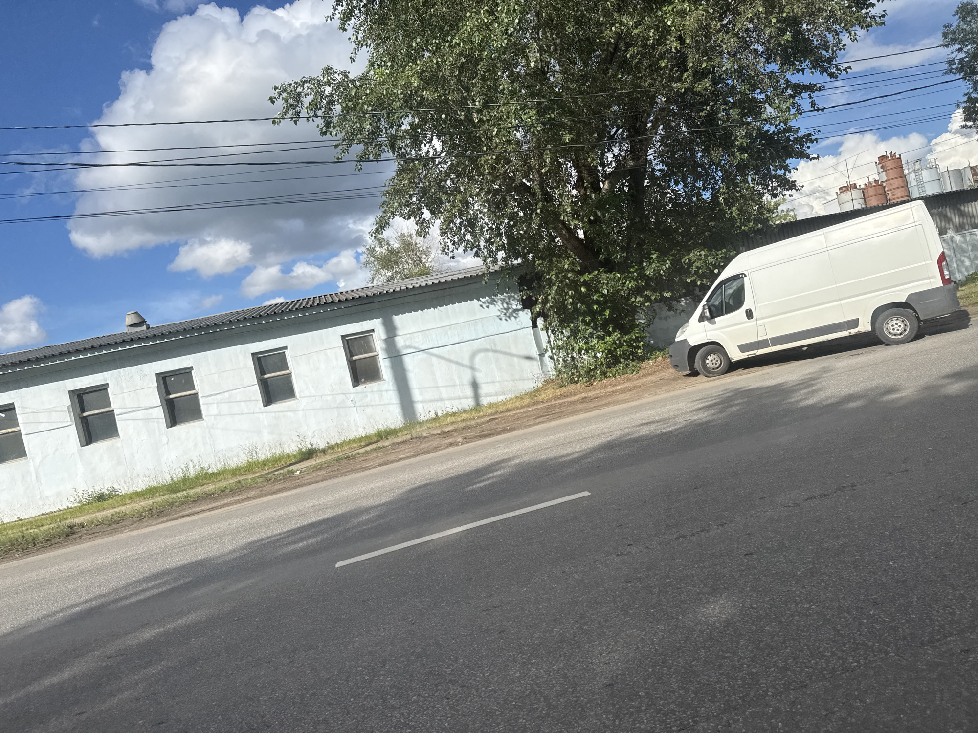 An outdoor, eye-level, medium shot captures a light blue, single-story building with a gray, corrugated roof running horizontally across the frame. The building has several dark-framed windows spaced evenly along its facade. In front of the building, a white panel van is parked on an asphalt road. The road occupies the lower half of the image and features a white line marking its edge. To the right of the van, a chain-link fence is visible, and beyond that, a row of industrial tanks with rusty-red tops. A large tree with dense green foliage is situated behind the building and to the left of the van, casting shadows on the road. The sky above is blue with scattered white clouds, and several black electrical wires are strung across the upper portion of the image. The scene is situated in Khimki, Russia.
