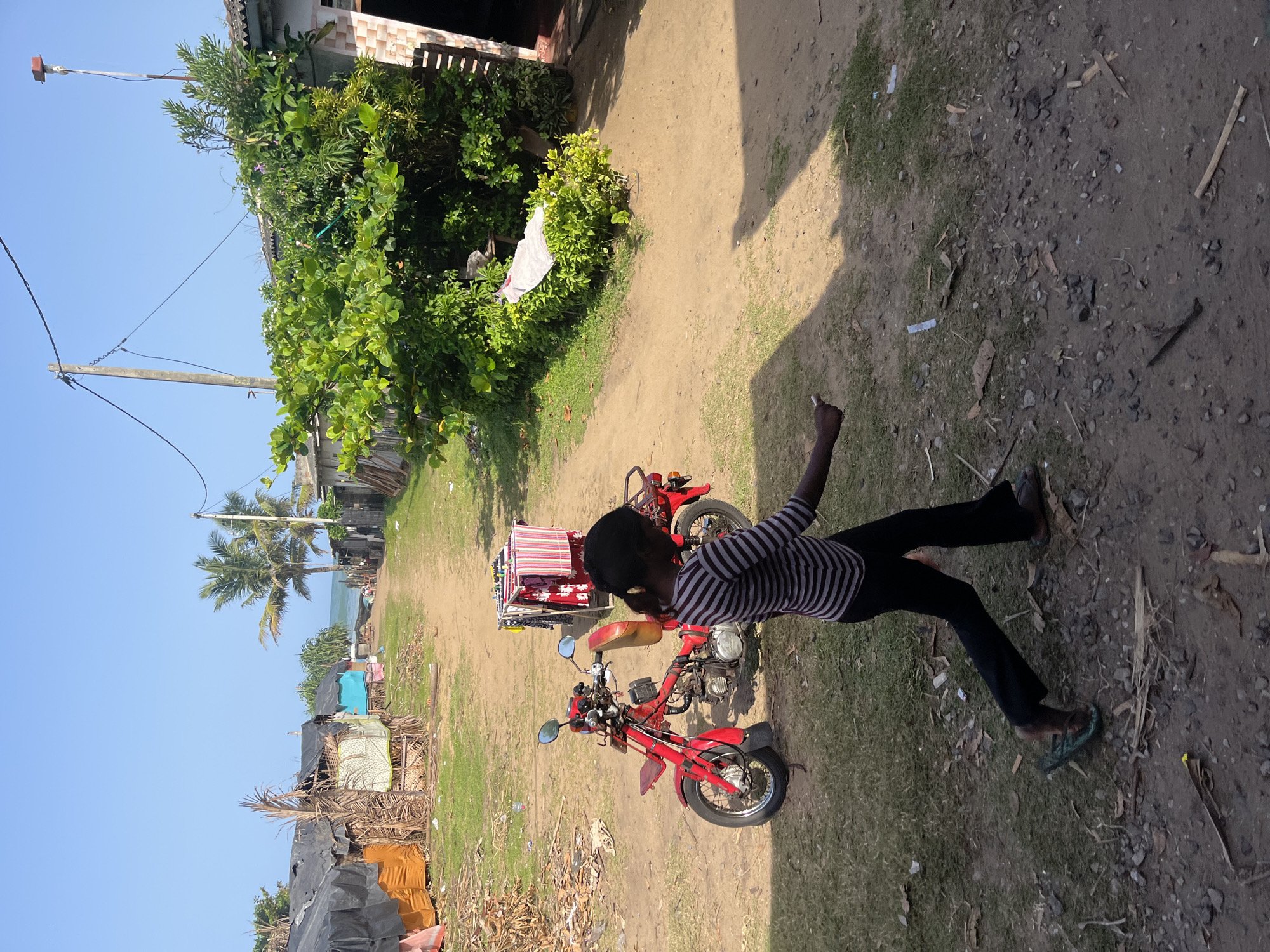 In Moratuwa, Sri Lanka, a young girl with dark hair, wearing a striped long-sleeved shirt and dark pants, walks along a dirt path lined with simple dwellings. To her left, a bright red motorcycle is parked. Behind the motorcycle, a clothes drying rack holds colorful folded fabrics. Palm trees and lush green foliage are visible in the background, with a glimpse of the ocean beyond. The environment appears to be a coastal community, with some structures showing signs of temporary construction and the ground littered with debris. The lighting suggests a sunny day.