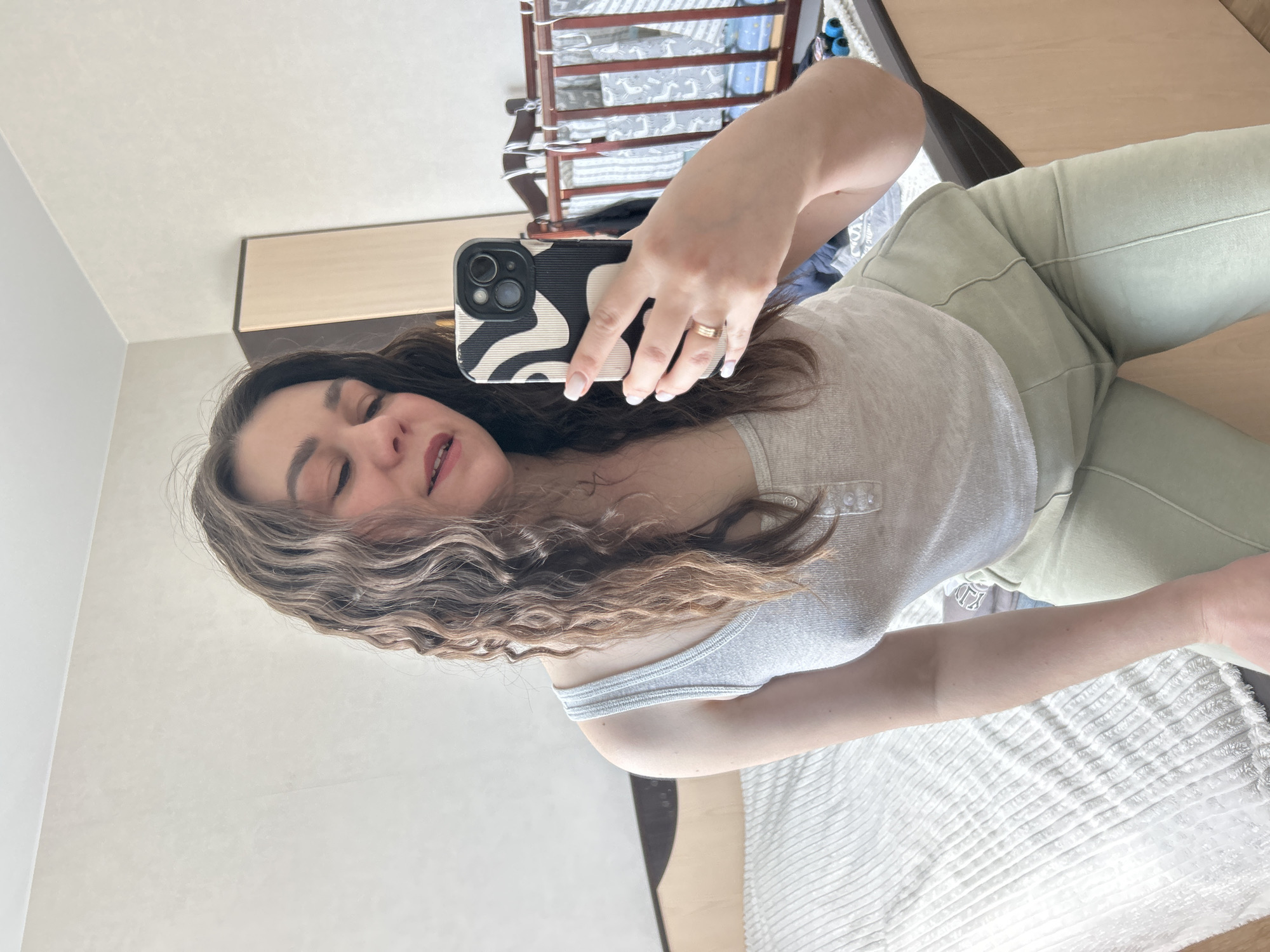 A woman is taking a selfie in what appears to be a bedroom. She is holding a phone with a black and white patterned case in her right hand. She is wearing a light-colored tank top and light green pants. Her hair is long and curly. The background shows a bed with a white comforter, a wardrobe, and a crib. The location is Chaikovsky, Russia.