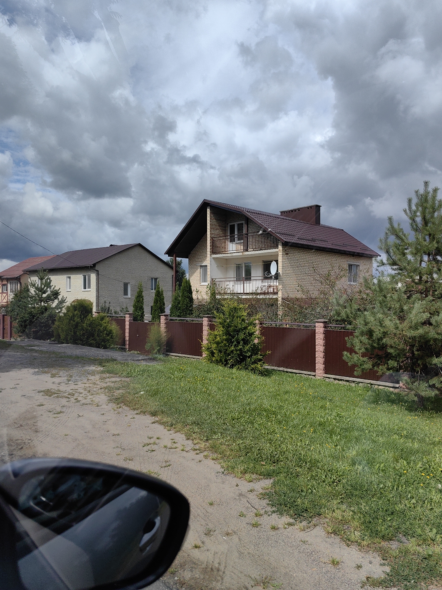 A daytime, eye-level, exterior shot shows a view of houses and a cloudy sky in the city of Myadzyel, Belarus. The foreground features the reflection of the surrounding area in a car's side mirror. Behind the mirror, a dirt road stretches into the distance, bordered by a strip of green grass on the right. A brown metal fence with brick pillars runs along the edge of the properties.
The main focus is on two houses. The house on the right is a two-story building with a brick facade, a dark brown pitched roof, and a prominent chimney. It has balconies on the upper level and windows on both floors. A satellite dish is visible on the side of the house. Next to it, a large green pine tree stands tall.
To the left of this house is another building, appearing to be two or three stories, with a light-colored textured exterior and a dark brown pitched roof. This house also has multiple windows and appears to be attached to or adjacent to another structure with a darker facade and a prominent cross-braced element. Various evergreen bushes and trees are planted in front of both houses, creating a landscaped appearance. The sky above is filled with dark, heavy clouds, suggesting overcast or impending weather.